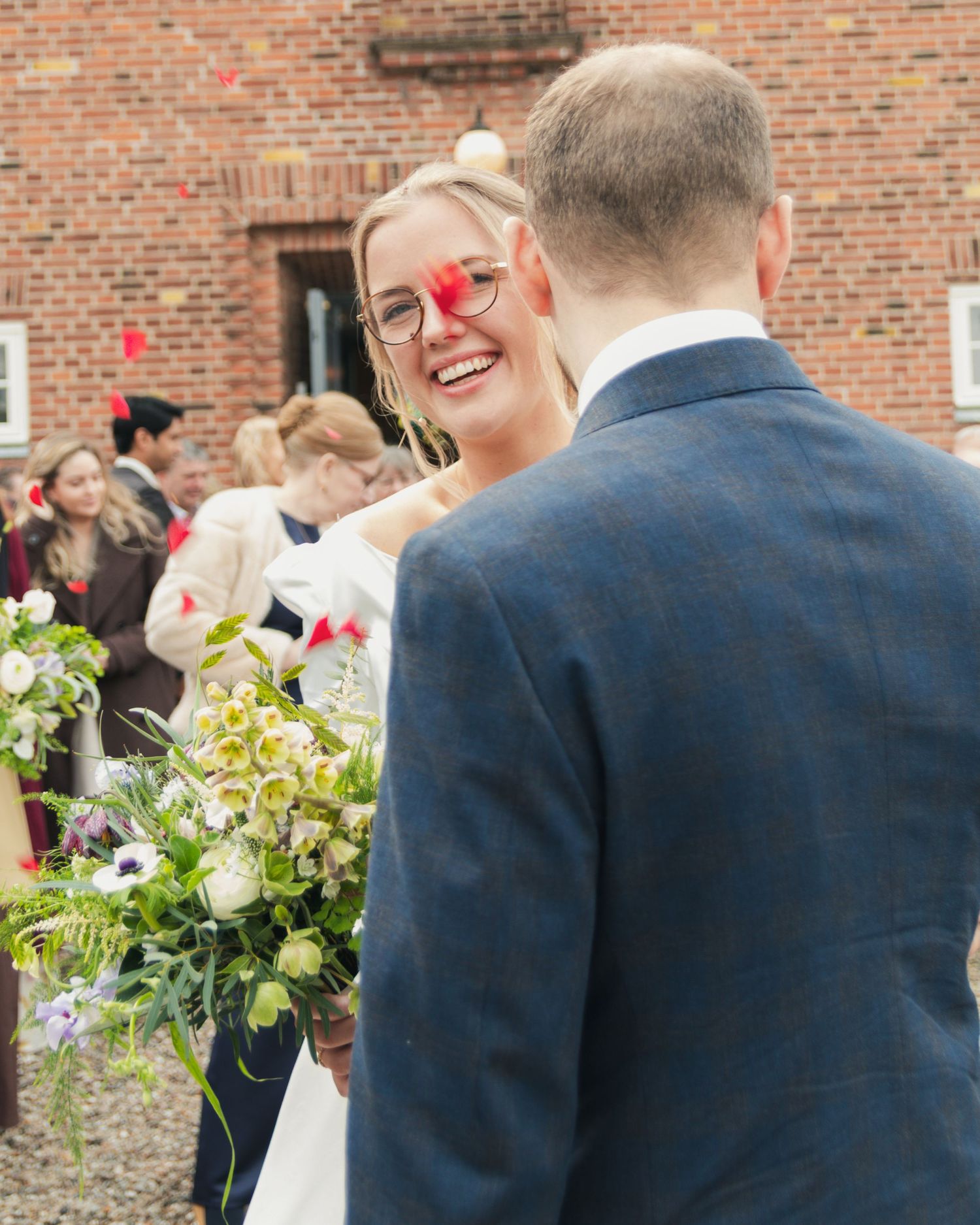 A laughing bride with a red petal on her face stands before her groom outside a brick church while guests celebrate behind them.