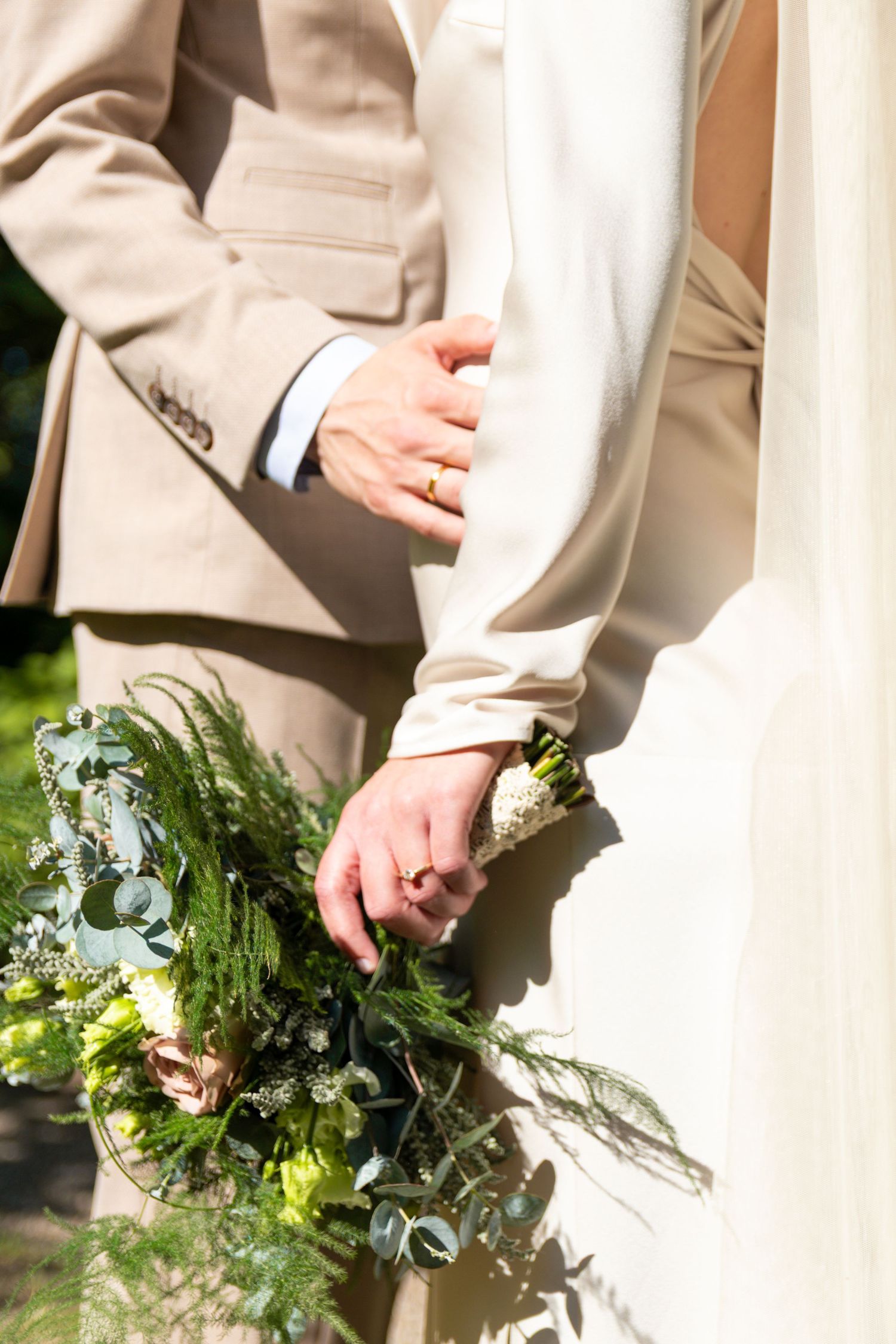 Close-up of a bride and groom standing together, showing wedding rings and a green bouquet of ferns and white flowers.