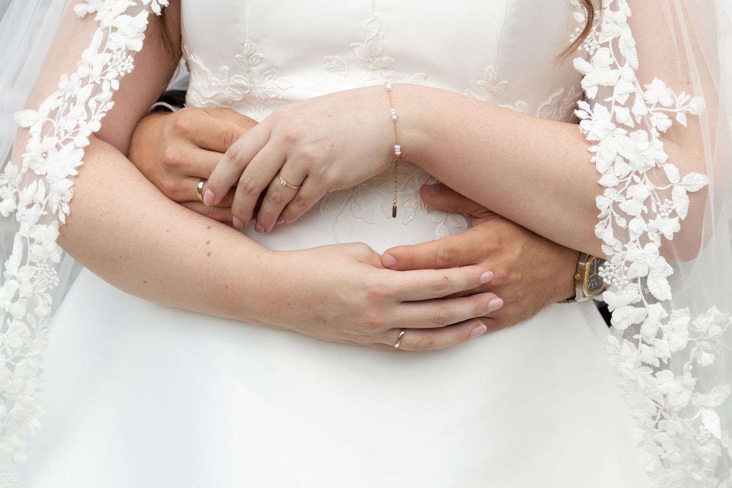 Close-up of a wedding couple's intertwined hands at the bride's waist, with lace veil trim framing her white dress.
