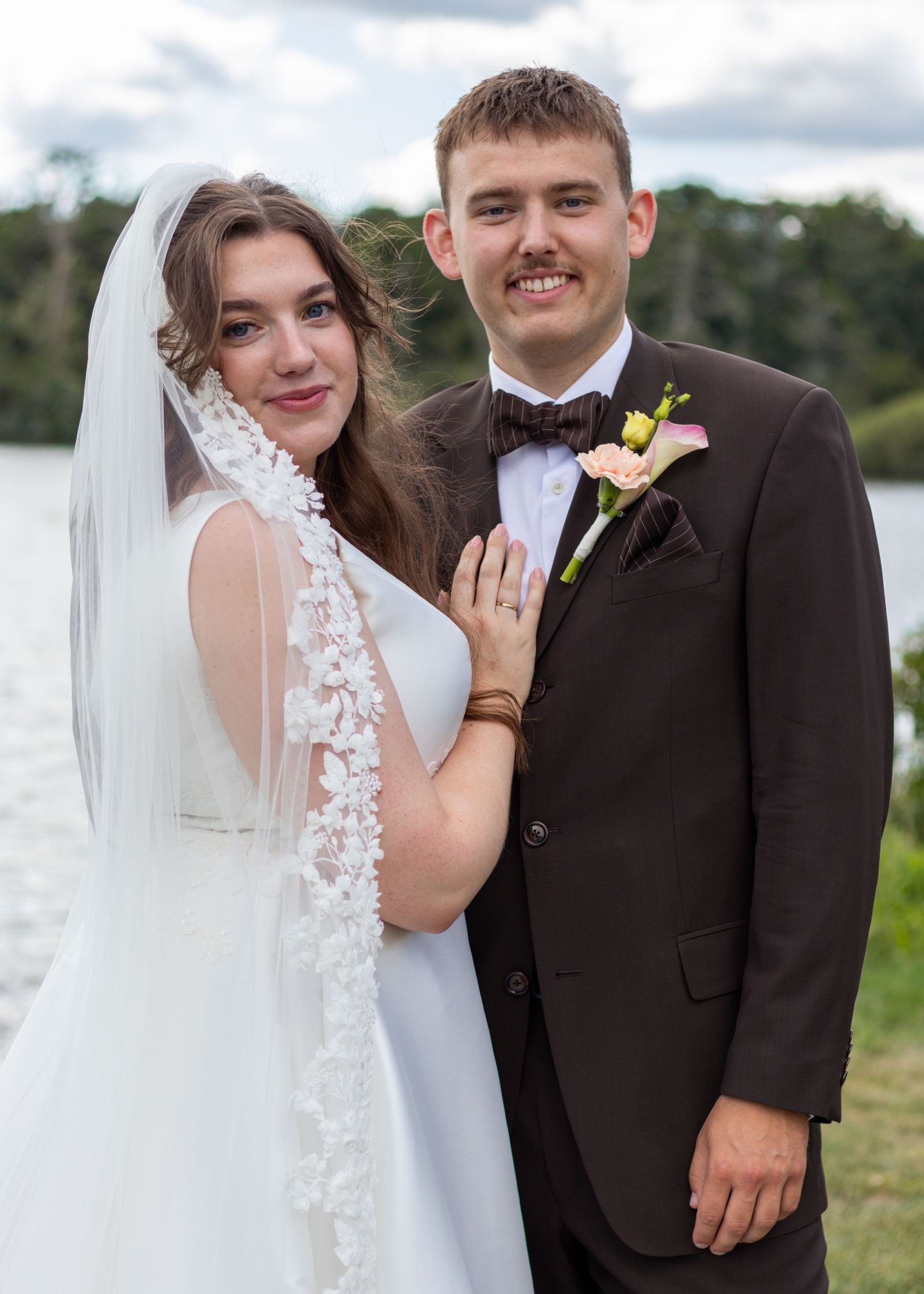 Smiling bride in a floral lace veil and groom in a brown suit with bow tie pose together by a lake.