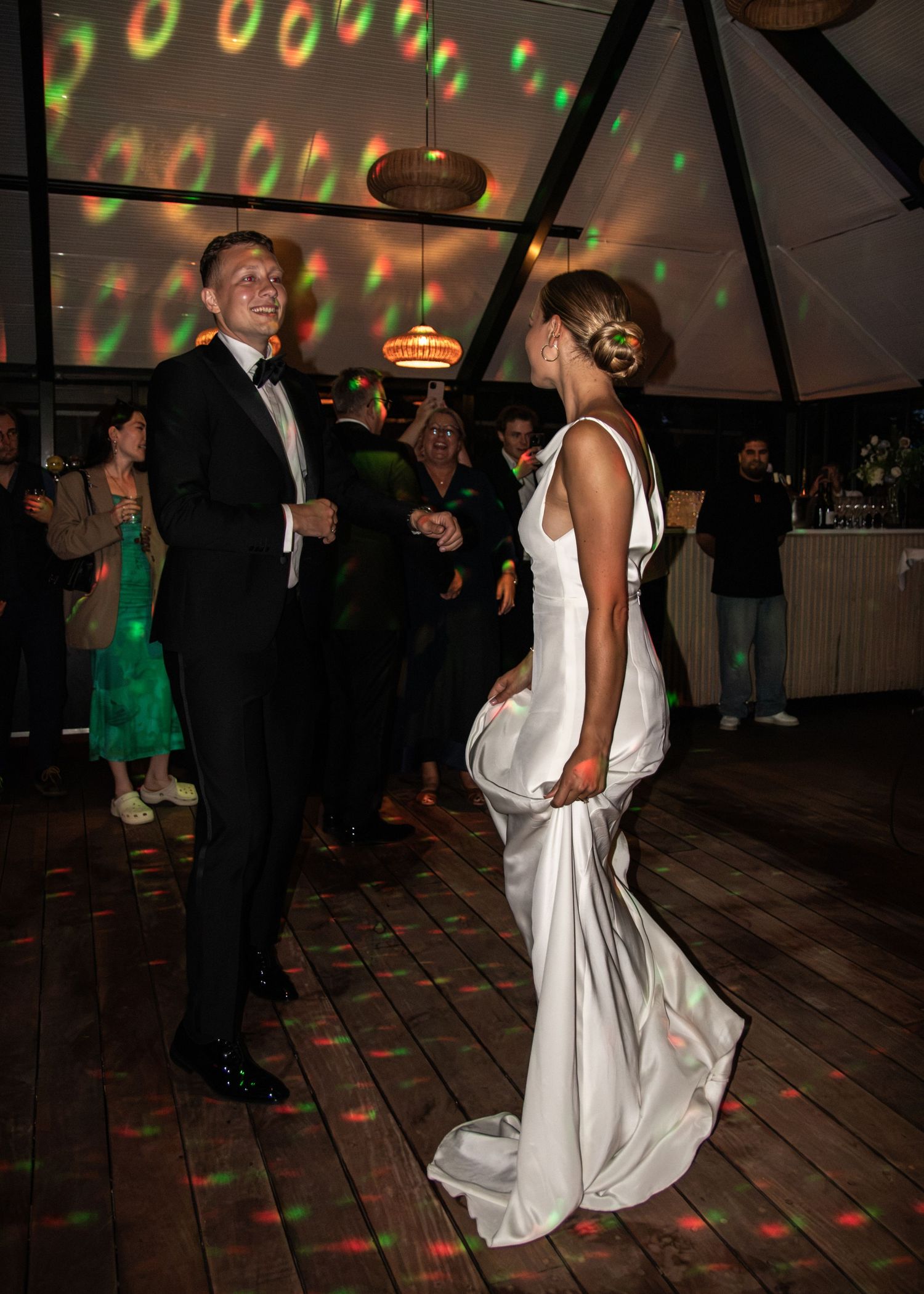 A groom in a tuxedo watches his bride in a white satin gown twirl on the dance floor lit by colorful party lights.