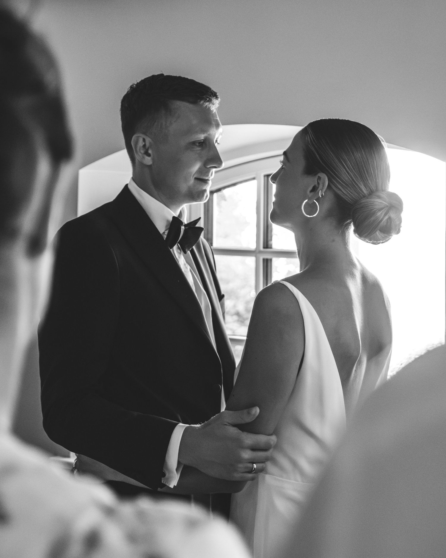 A black-and-white photo of a groom in a tuxedo gazing tenderly at his bride in a white dress beside an arched window.
