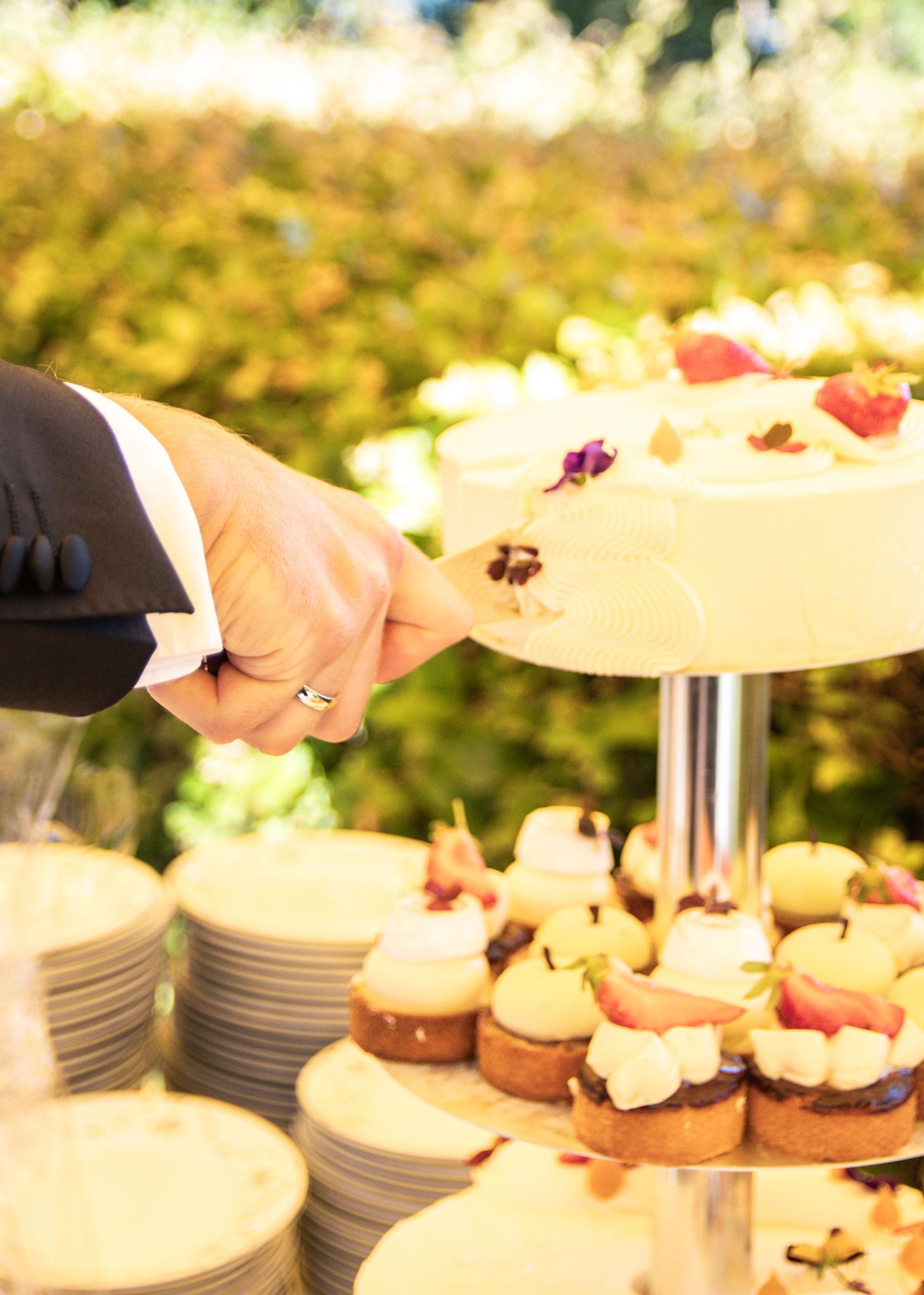 A hand with a ring reaches toward a tiered stand of wedding cake and fruit-topped mini pastries in a garden setting.