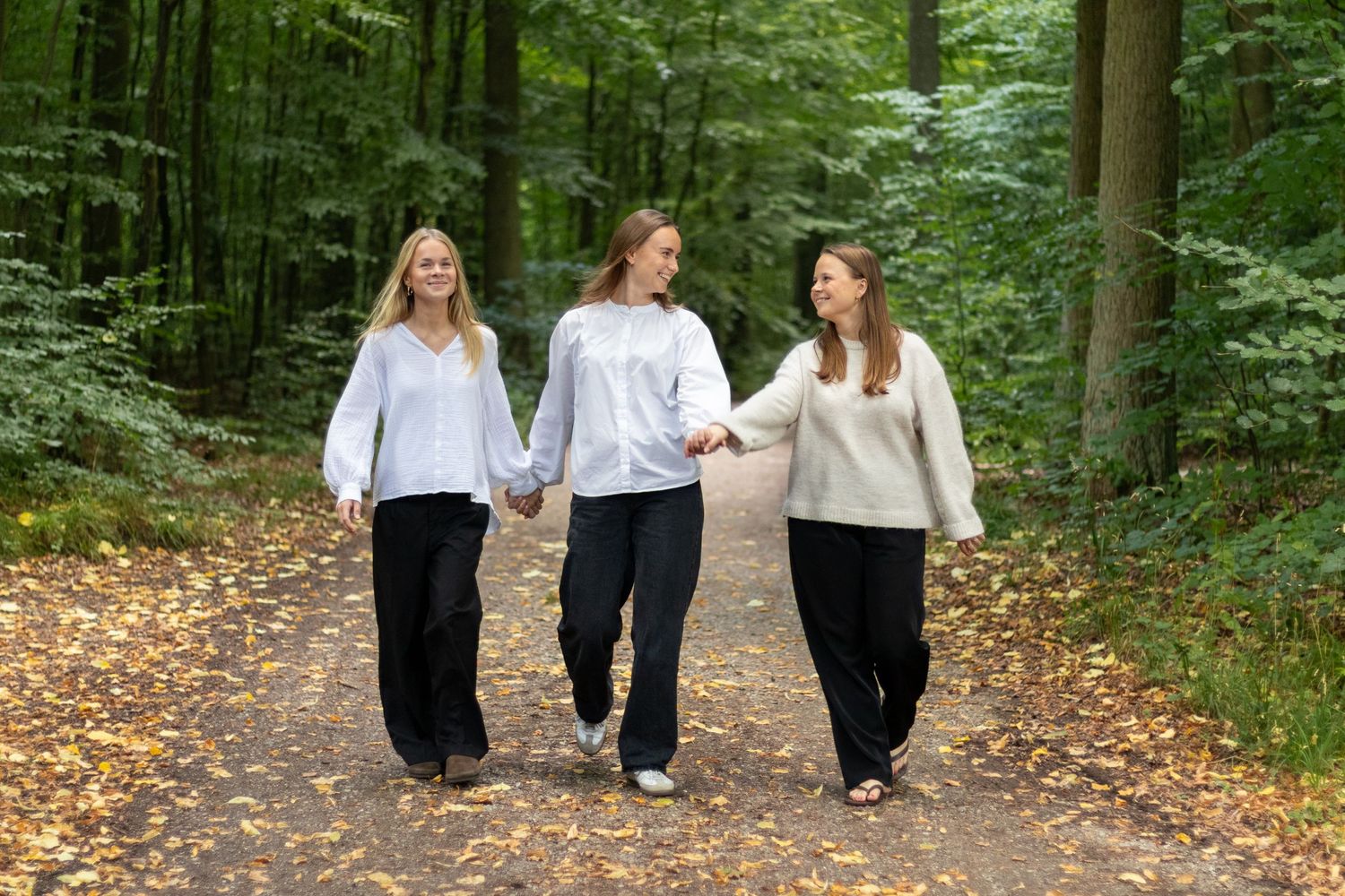 Three young women in white tops and black pants hold hands and walk together along a leaf-covered forest path.