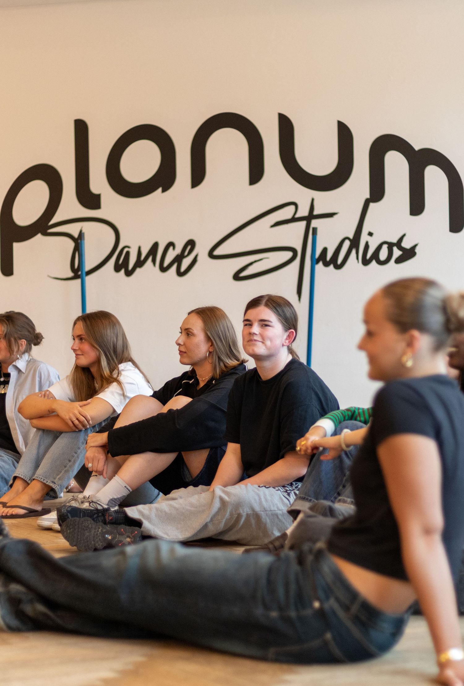 Young women sit on the floor in a row beneath a Planum Dance Studios wall sign, listening attentively during a class.
