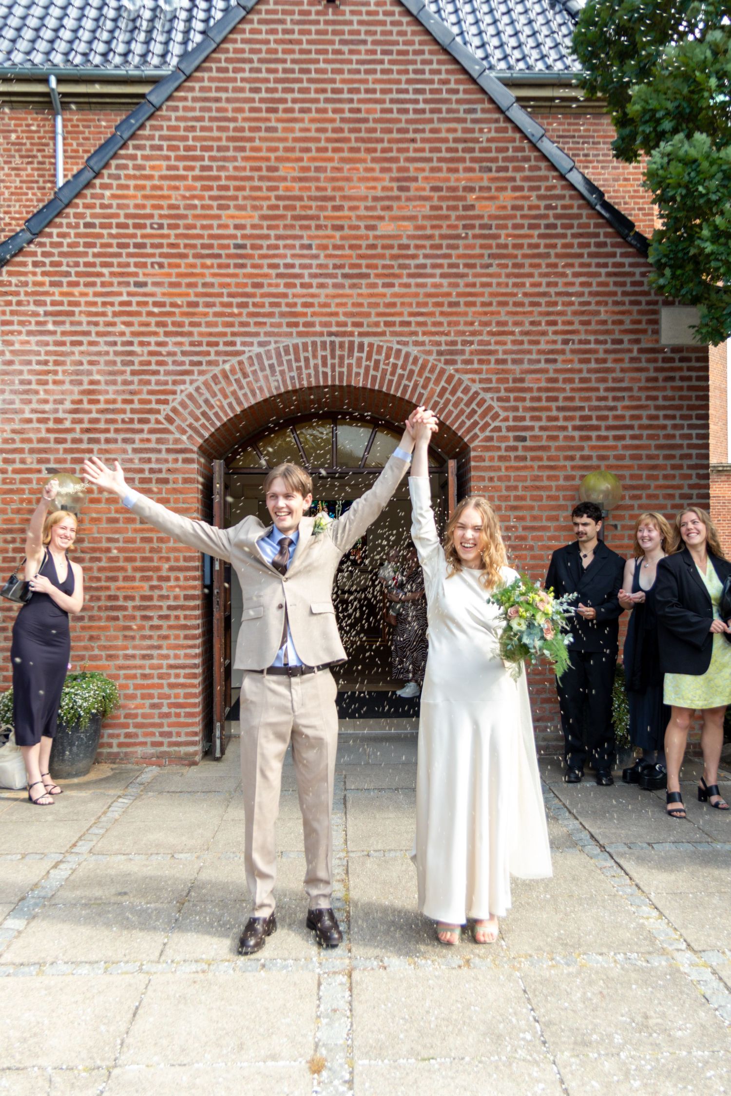 Newlywed couple in a beige suit and white dress cheer with raised arms outside a brick church as guests watch.