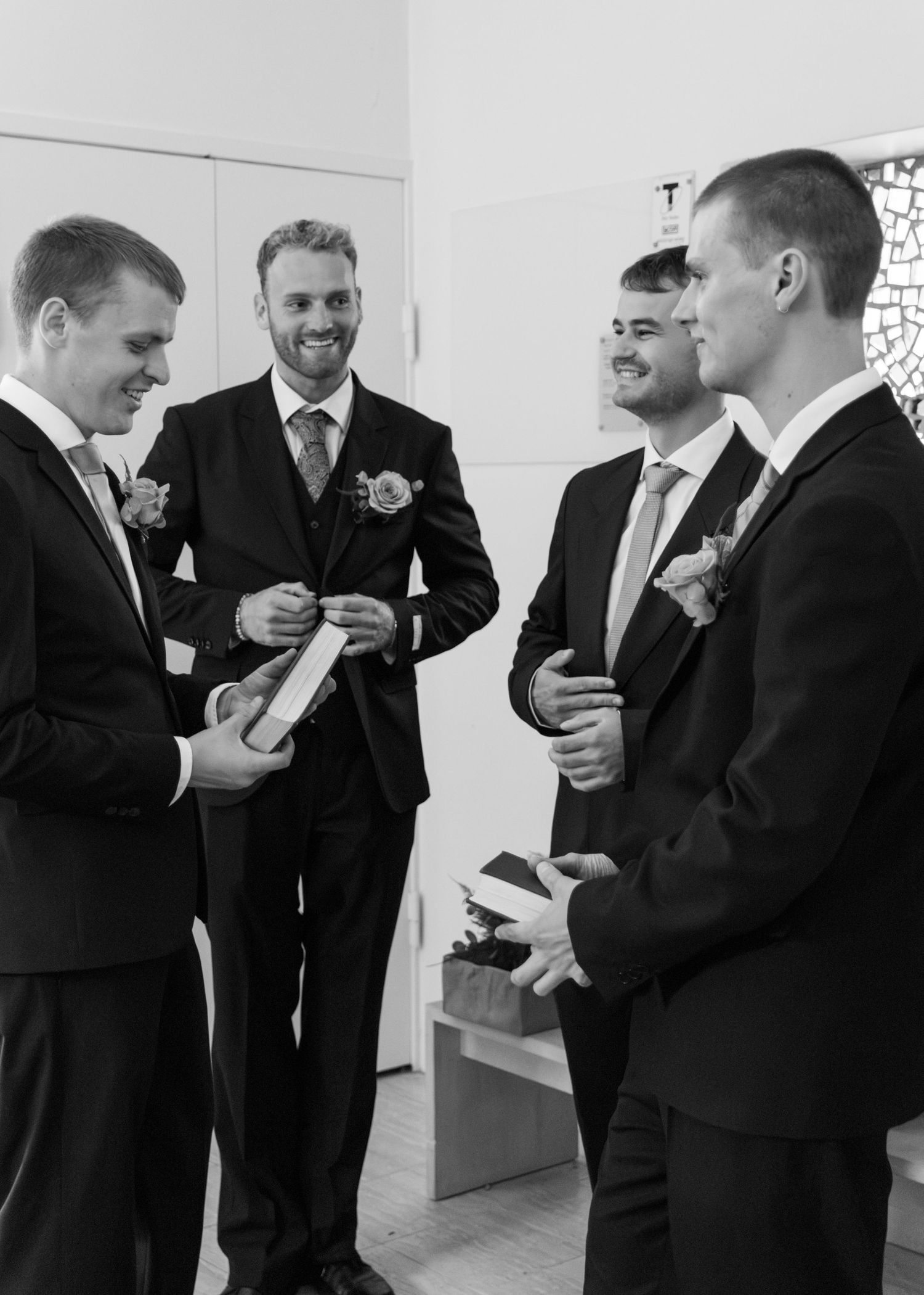 Four groomsmen in dark suits with boutonnieres smile and hold small gift boxes while chatting before the wedding.
