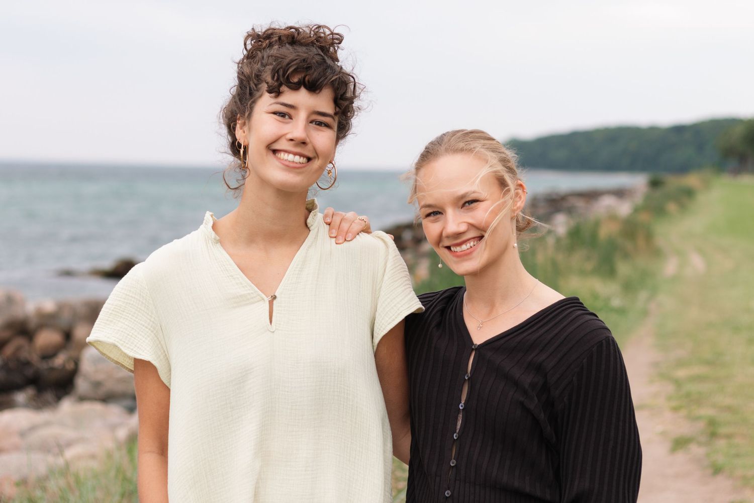 Two cheerful women pose close together on a shoreline trail with rocky beach, calm sea and green hills in the background.