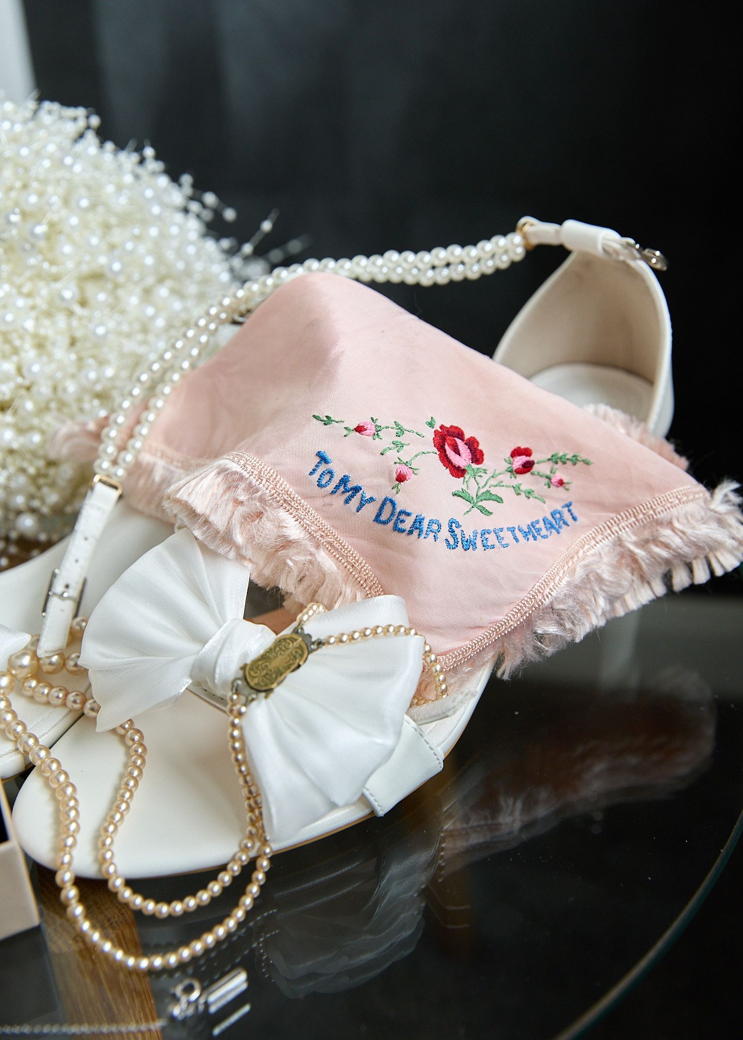 White bridal heels with bow detail beside a pink embroidered handkerchief reading To My Dear Sweetheart, surrounded by pearl necklaces.