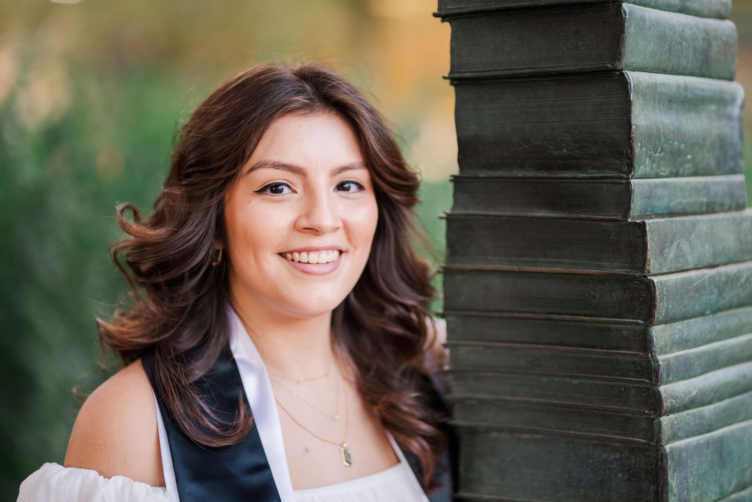 Young woman with long brown wavy hair smiling beside a green pillar outdoors in a portrait photo.