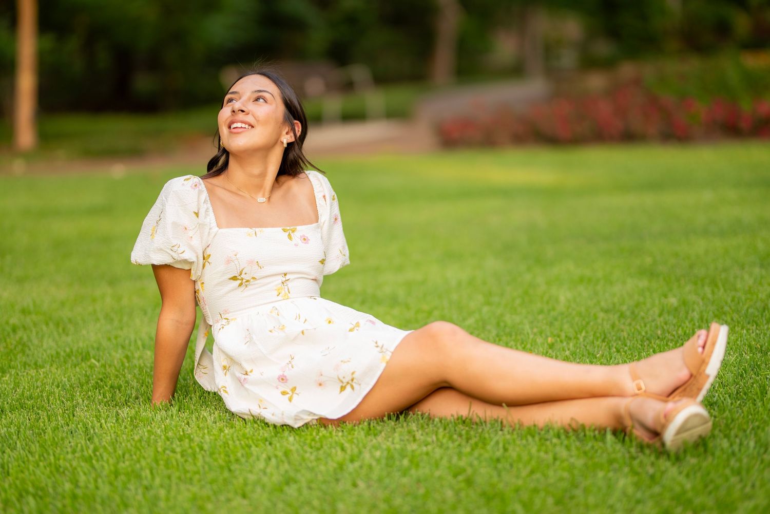 Young woman in white floral dress sitting on lush green grass in a park, smiling and looking up joyfully.