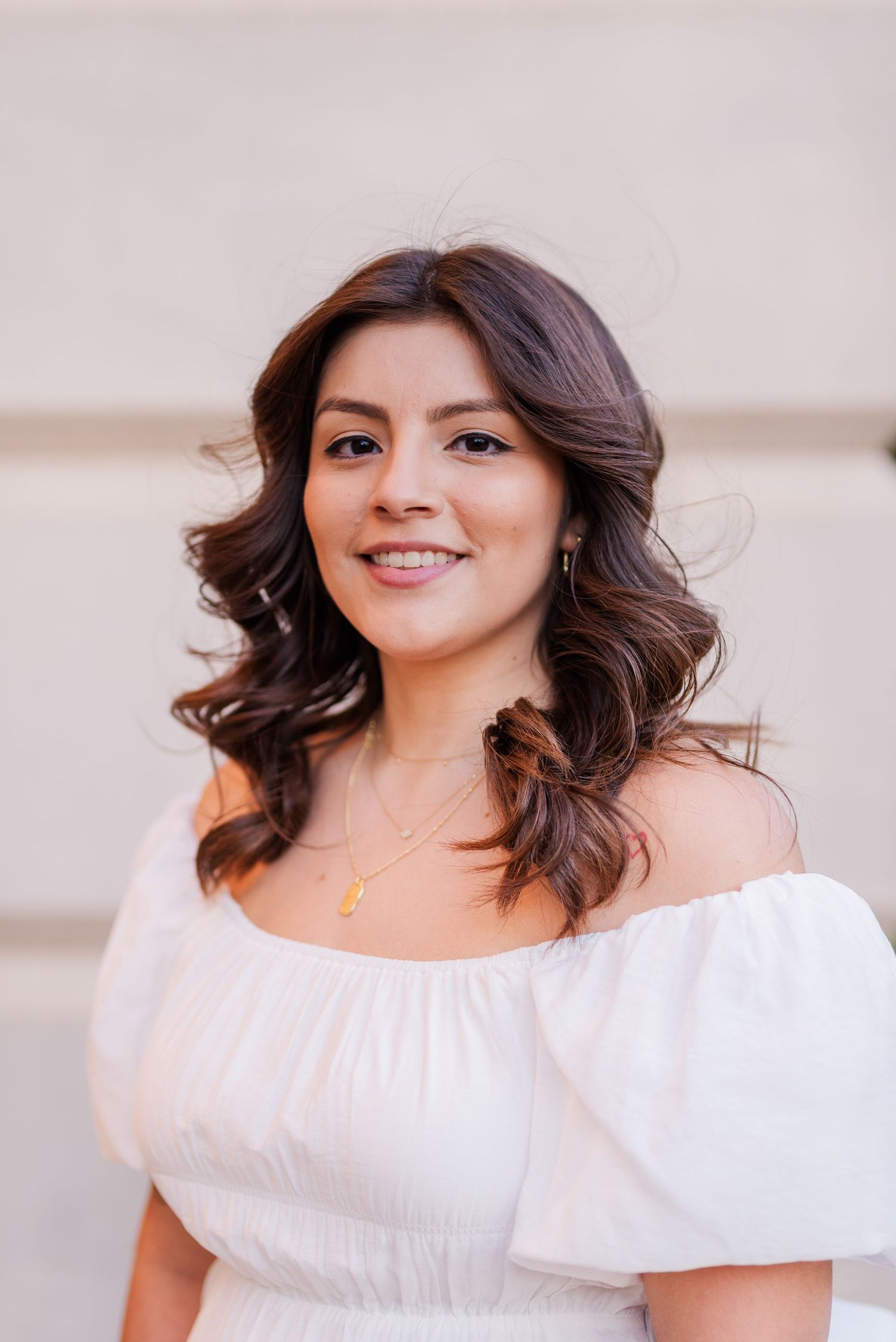Young woman in white off-shoulder dress with gold necklace, smiling outdoors against a neutral background.
