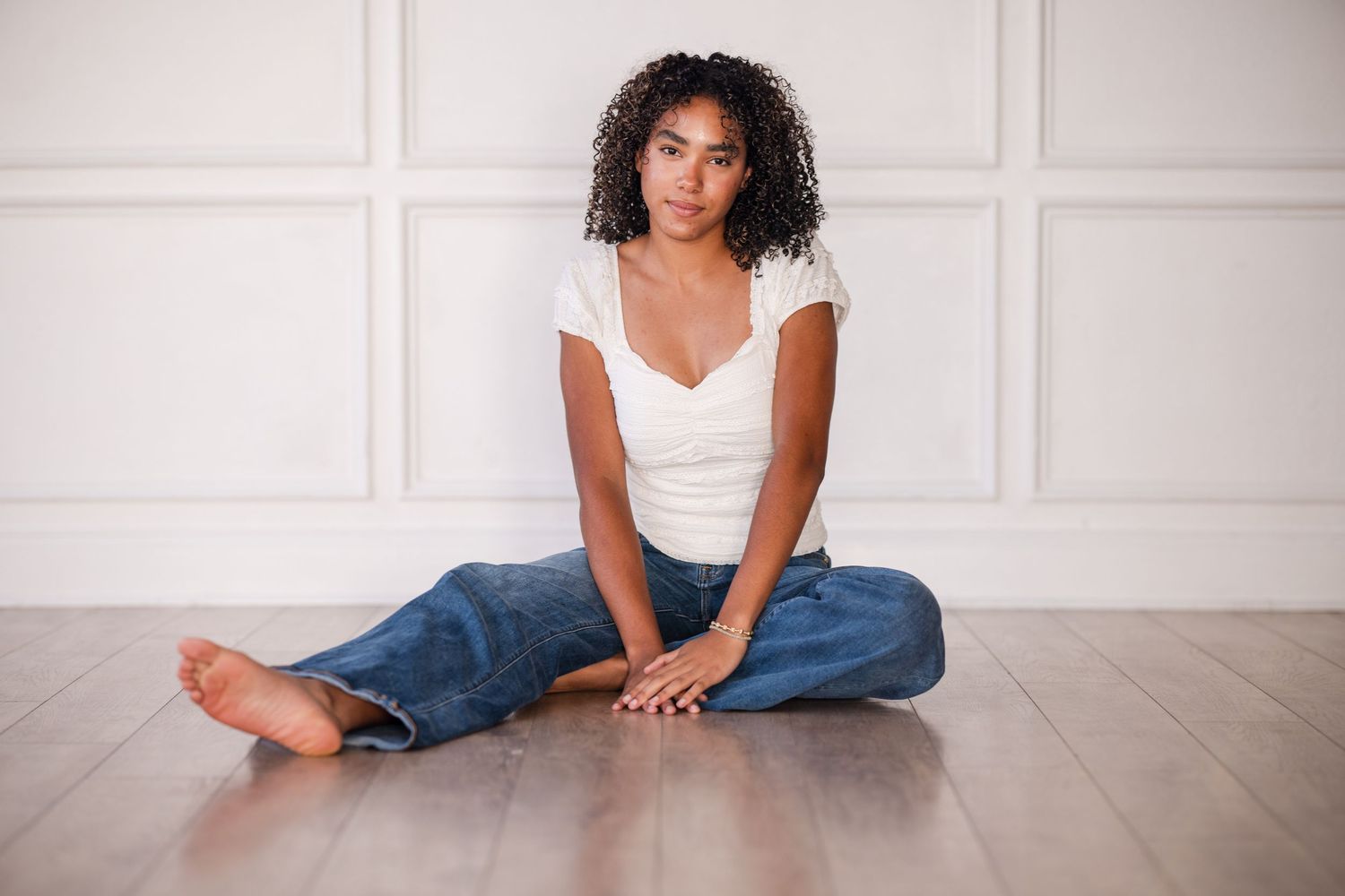 Young Black woman with curly hair sits cross-legged on hardwood floor wearing white top and blue jeans against white wall.