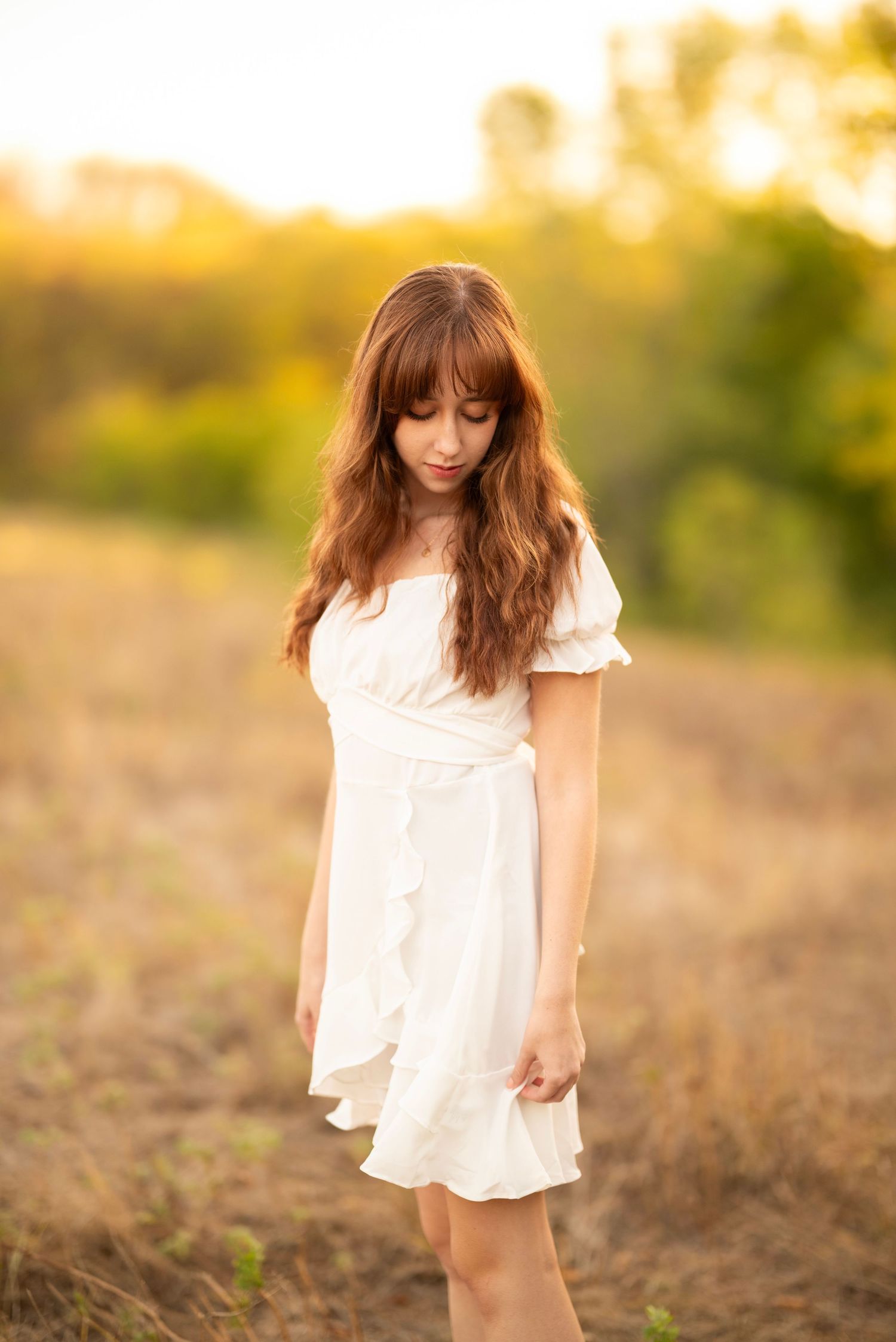 Girl standing in field for senior photos with beautiful warm lighting while wearing a white dress