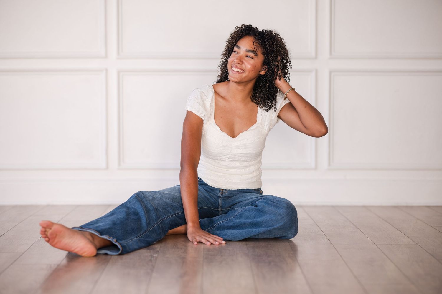 Young Black woman sitting cross-legged on hardwood floor, wearing white crop top and blue jeans, smiling cheerfully.