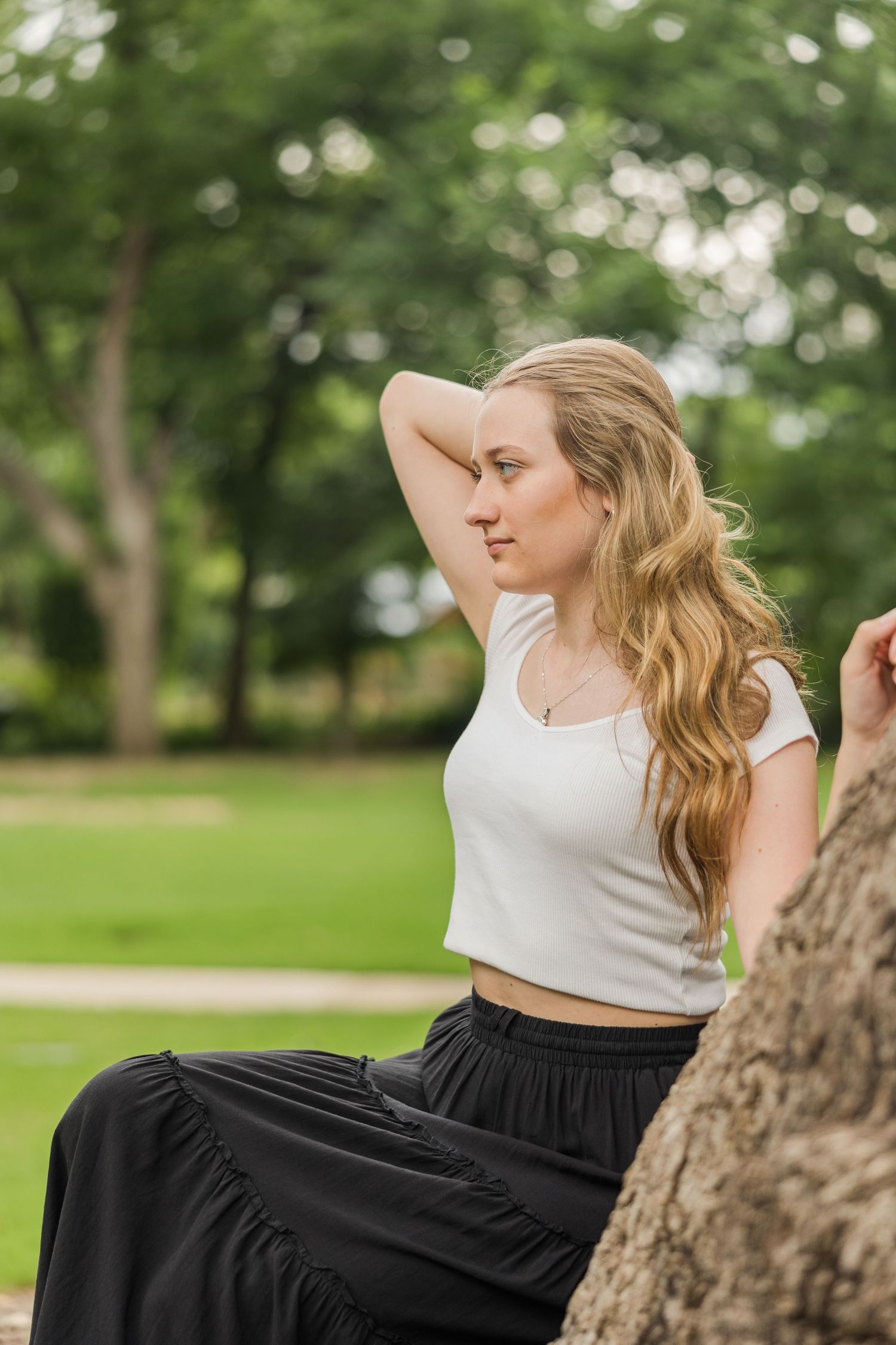 Young blonde woman in white crop top and black skirt sits by a tree in a lush green park.