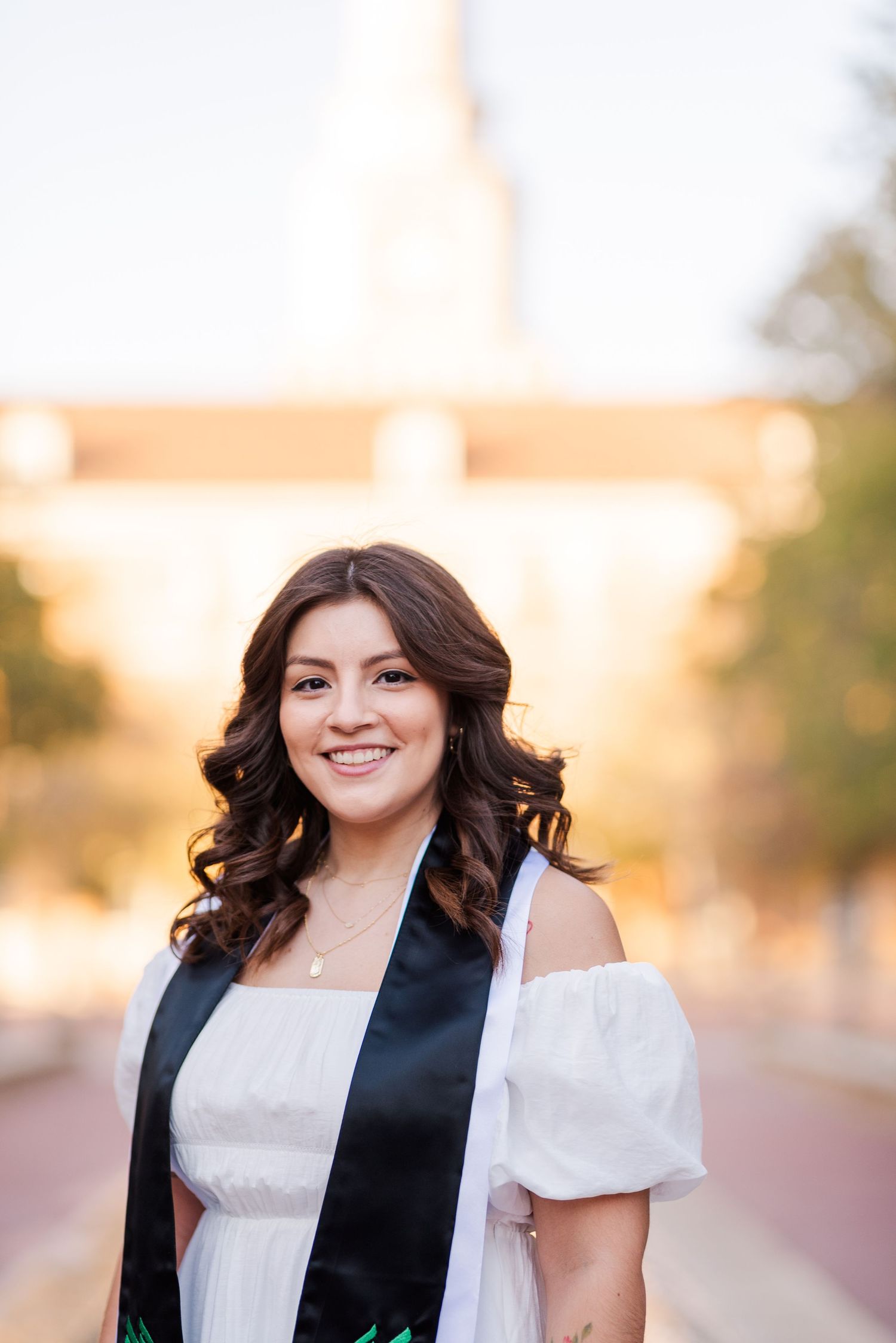 Smiling young woman in white dress with black graduation stole posing outdoors near a university building.