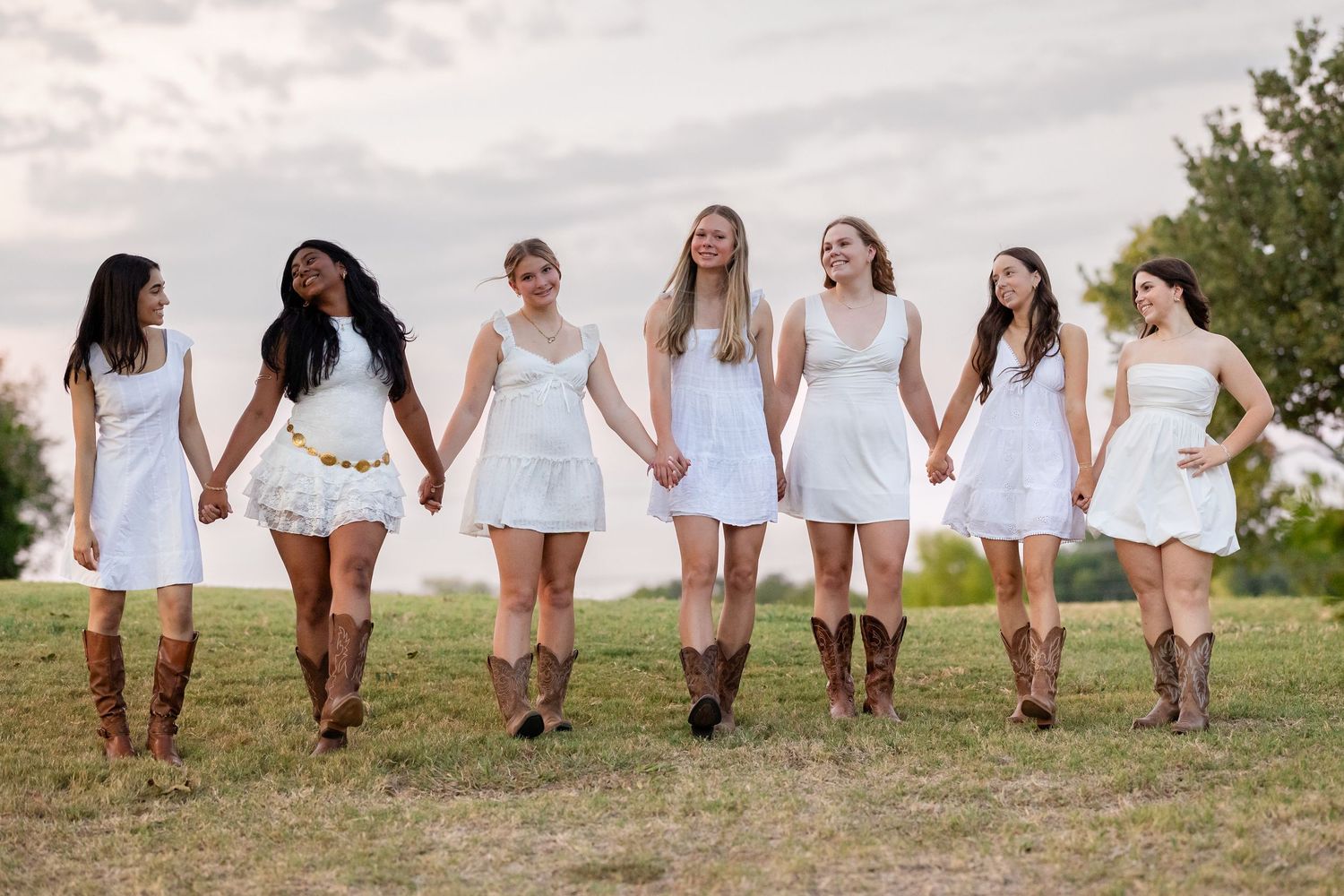Seven young women in white dresses and cowboy boots hold hands walking through a green field outdoors.