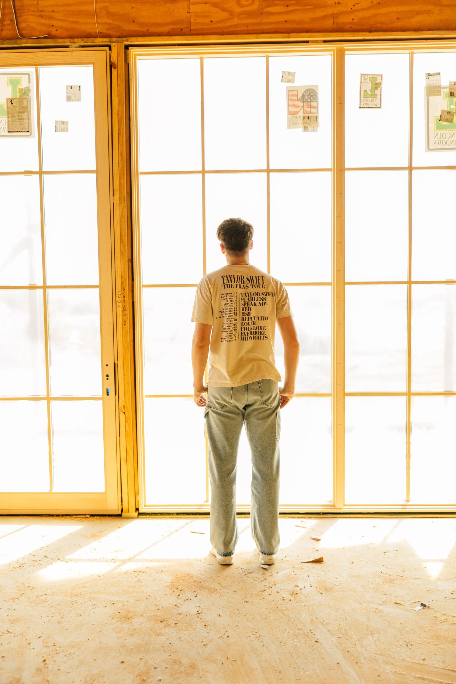 Man in graphic tee and gray pants stands facing large bright windows in a wood-floored room under renovation.