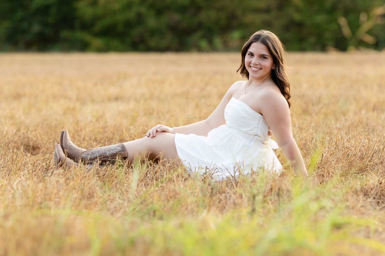 Young woman in white dress and cowboy boots sitting gracefully in a golden field during a sunny outdoor senior photo session.