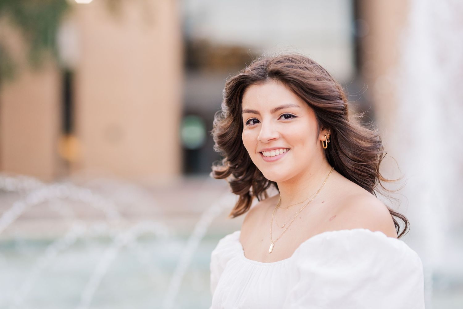 Young woman in white off-shoulder top smiling near a fountain, wearing a delicate necklace outdoors.