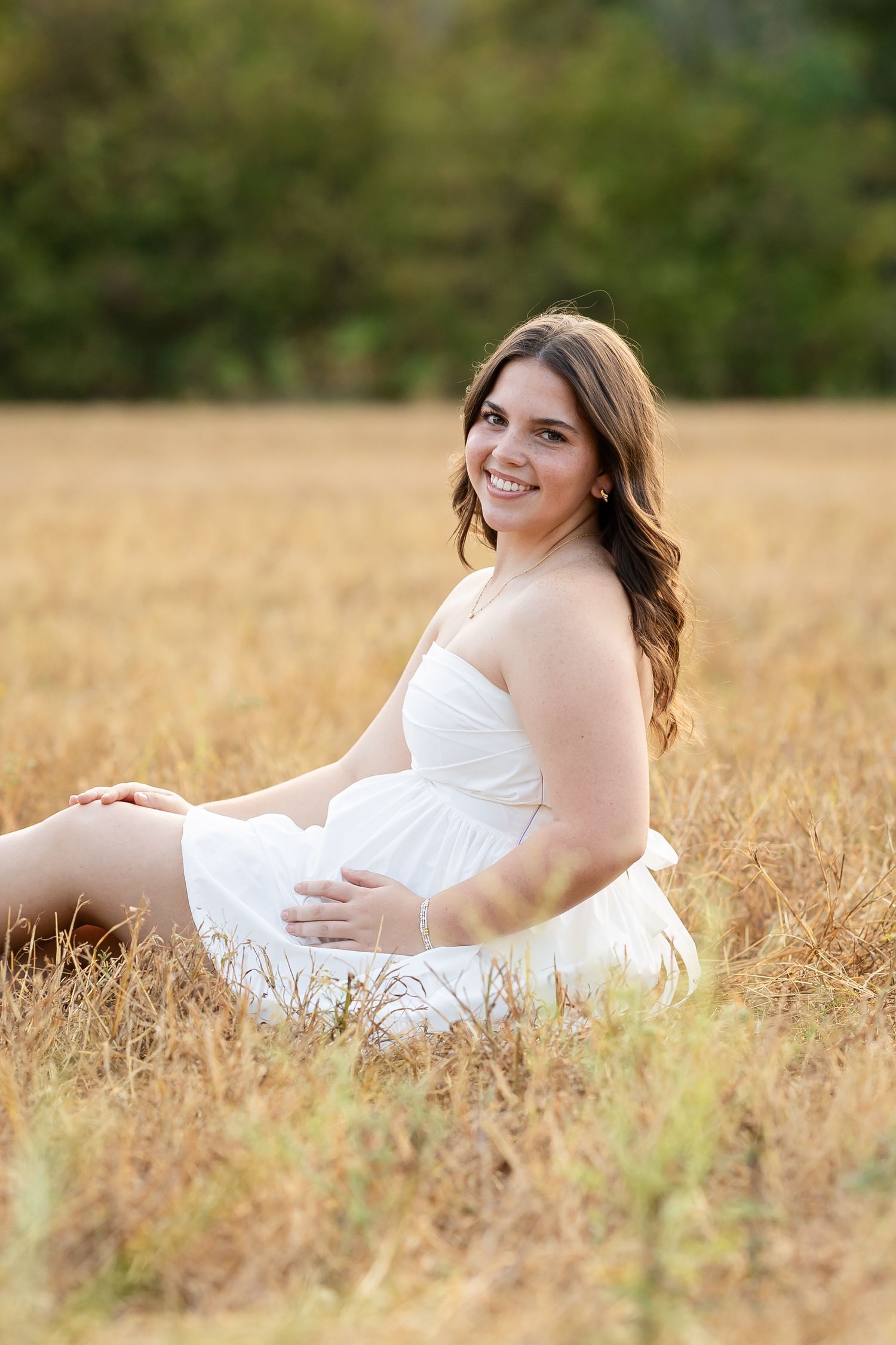 Young woman in white dress sitting in golden field, smiling outdoors with green trees in background.
