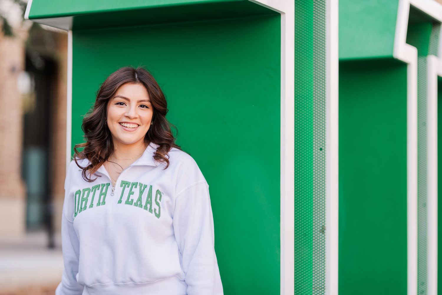Smiling young woman in a North Texas sweatshirt posing outdoors near a green structure on campus.