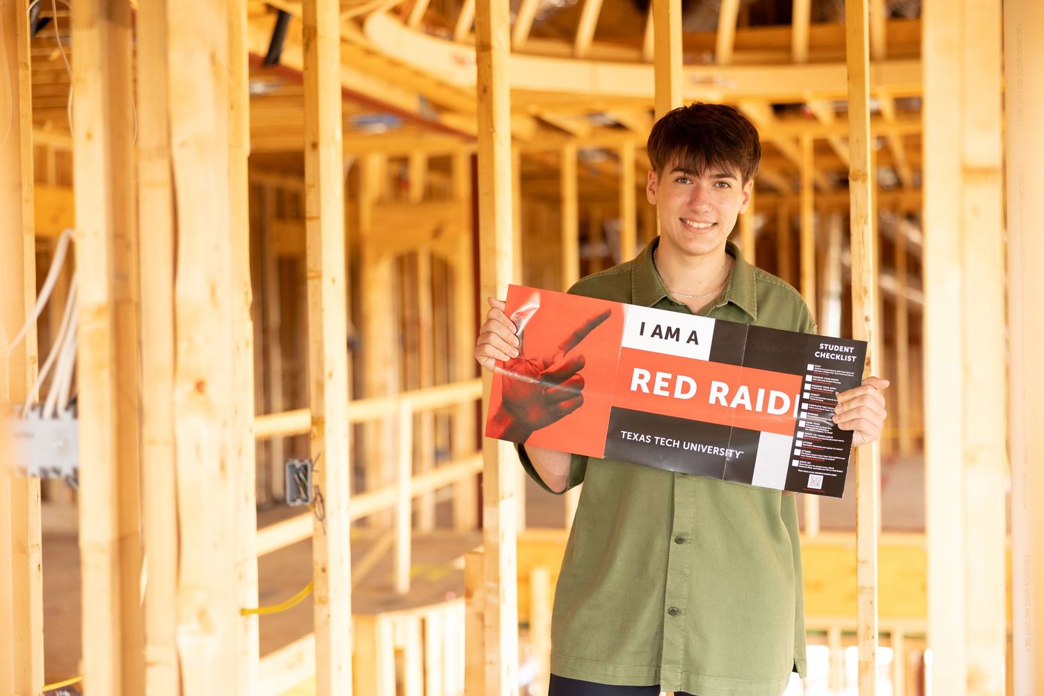 Young man holding a Red Raider Texas Tech University sign inside a wood-framed building under construction.