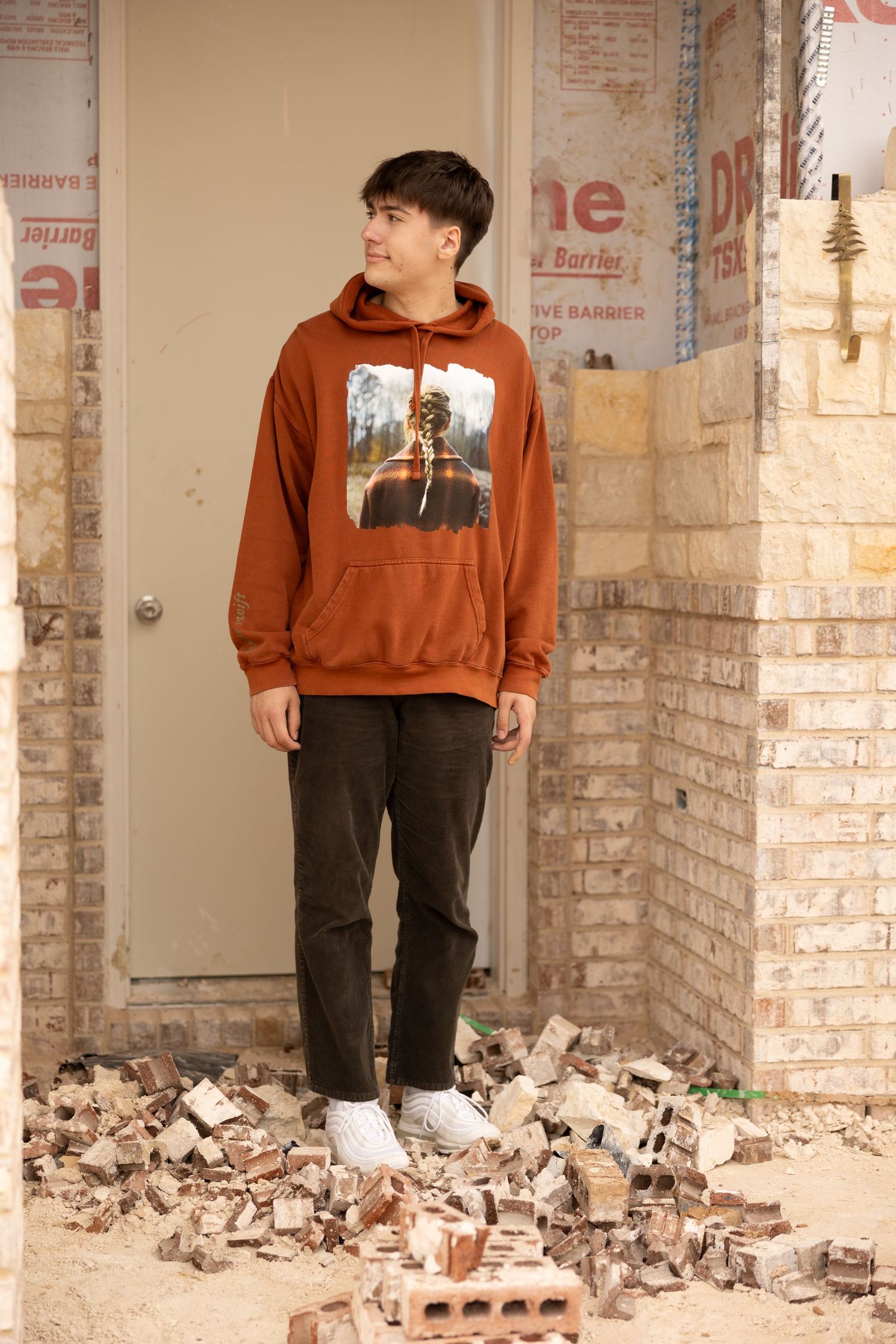 Young man in rust orange graphic hoodie and dark pants stands among scattered bricks at a construction site.