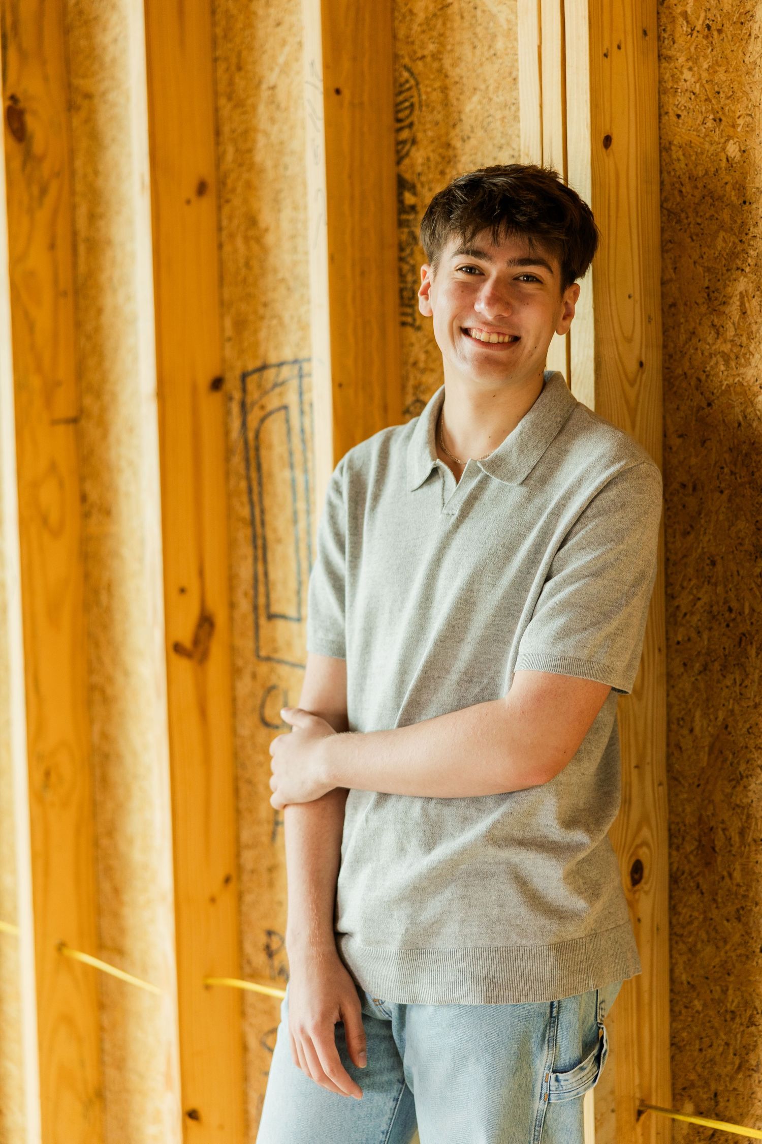 Smiling teenage boy in gray polo shirt leaning against wooden wall with arms crossed.