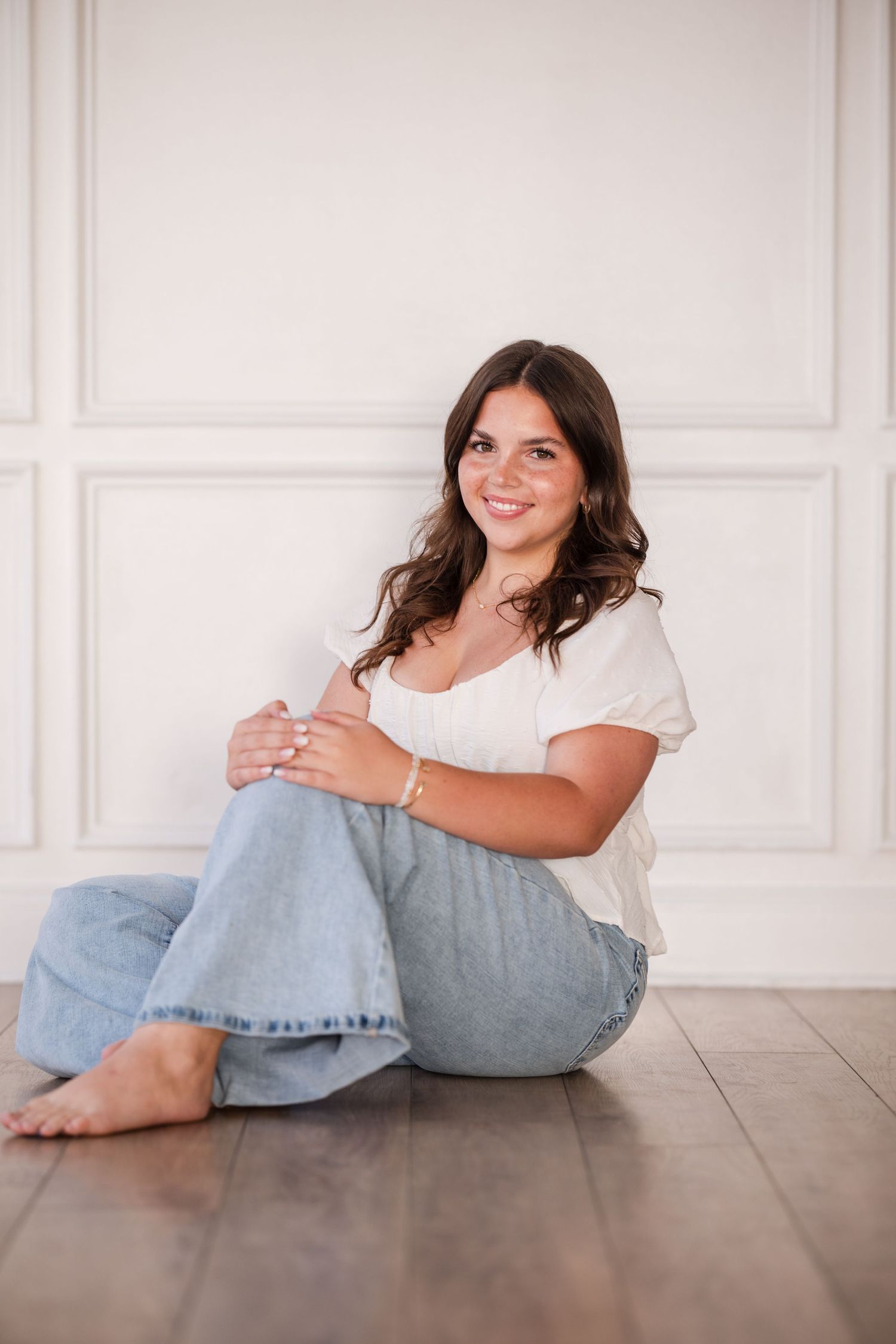 Young woman with dark hair sitting on hardwood floor against white wall, wearing white top and light blue jeans.