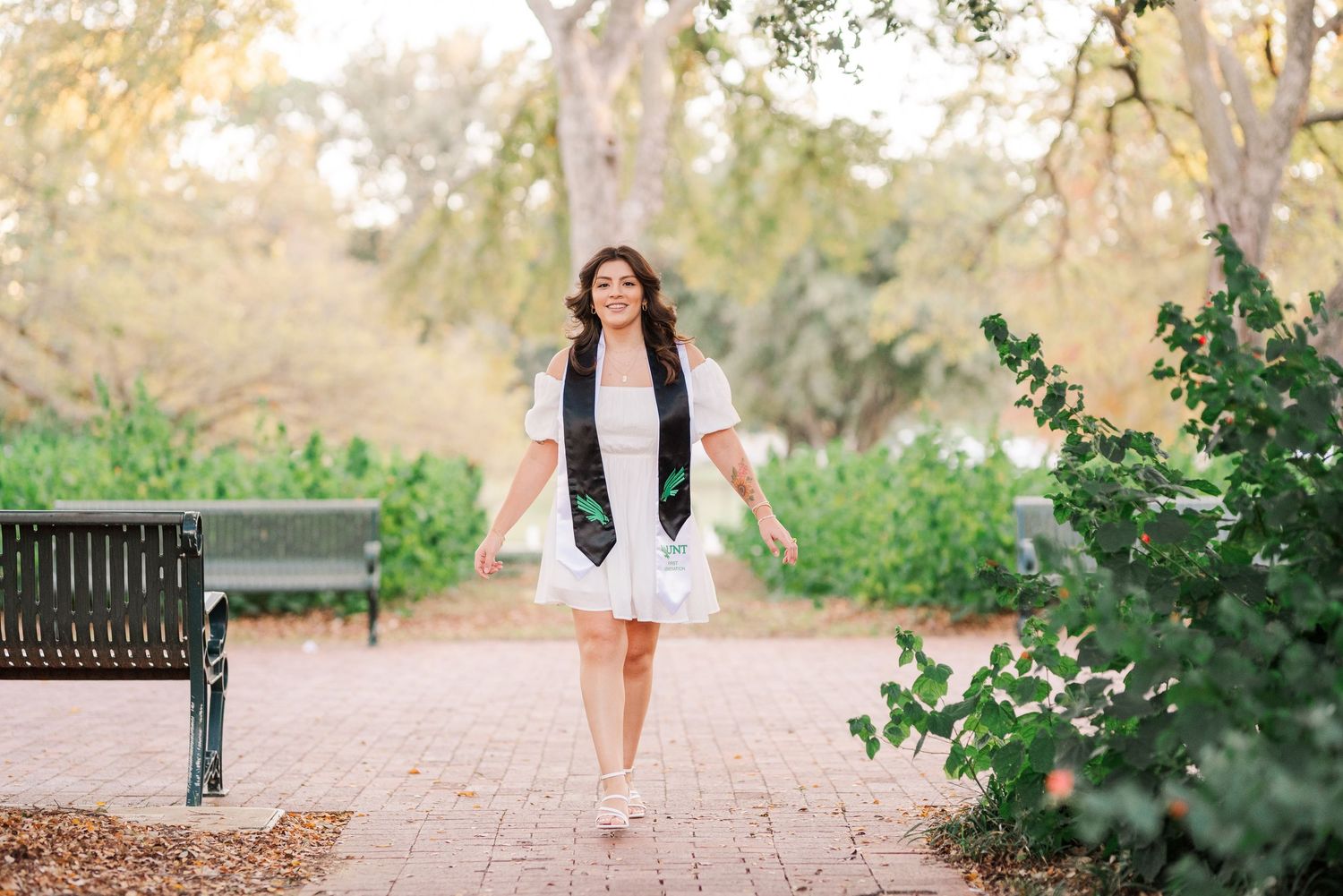 Young woman in white dress and graduation stole walking on brick path in park setting.
