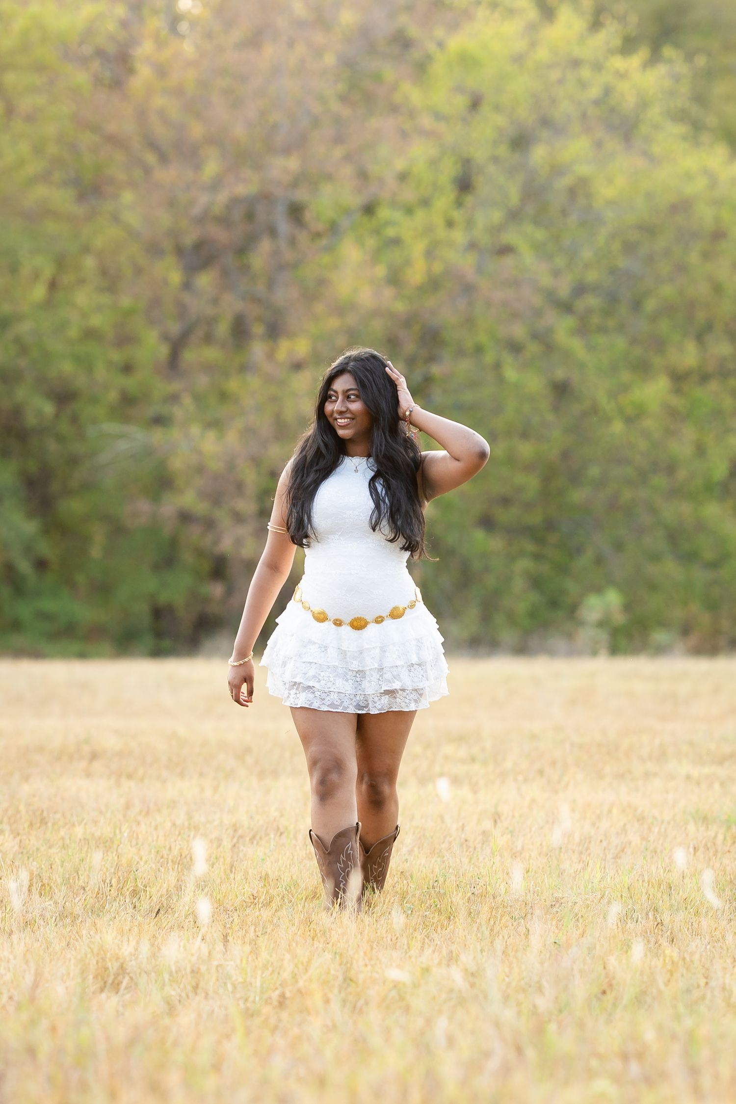 A smiling woman in a white ruffled mini dress, gold belt, and cowboy boots stands in a golden field.