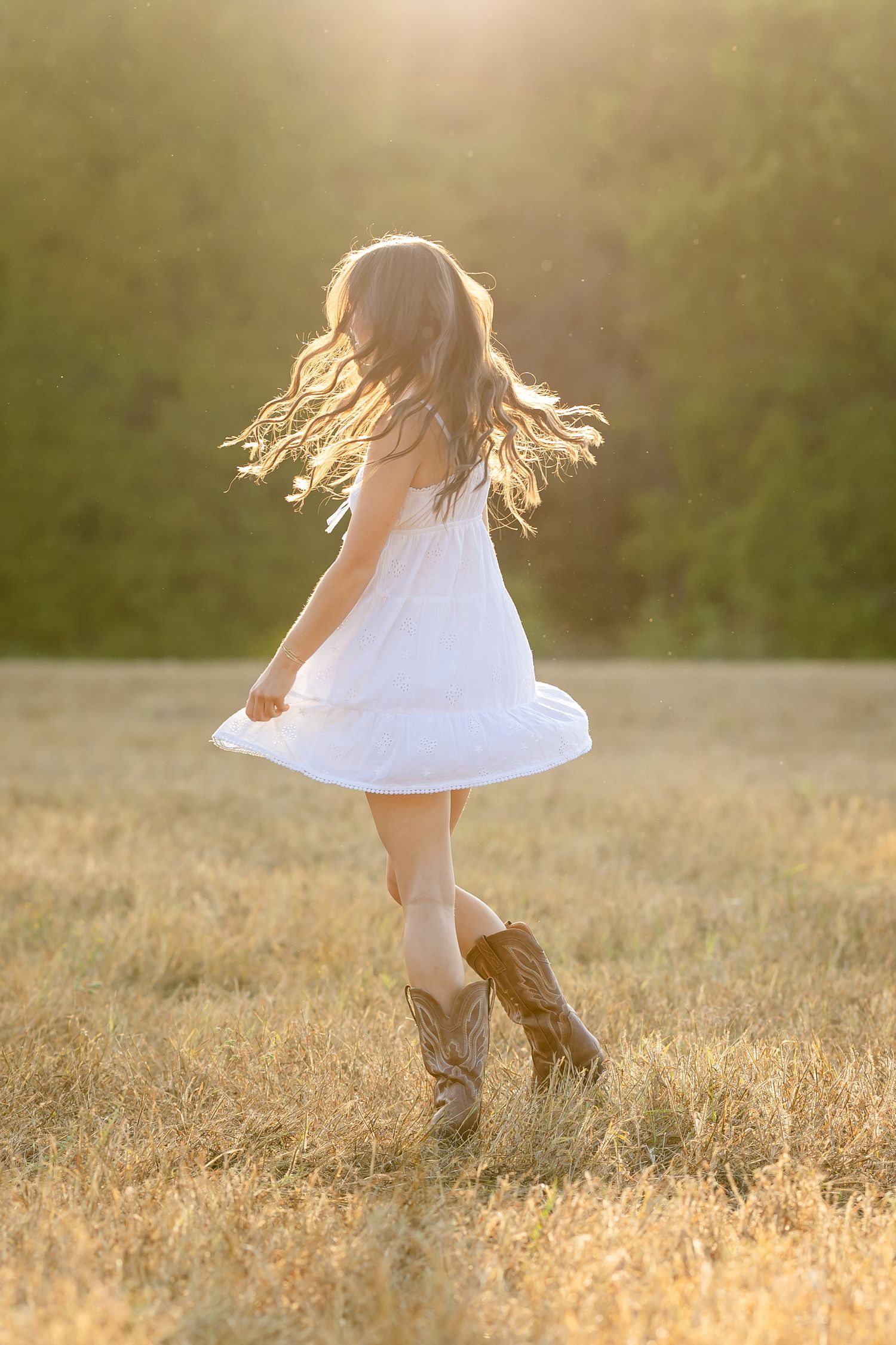 A young girl in a white dress and cowboy boots twirls joyfully in a golden sunlit field.