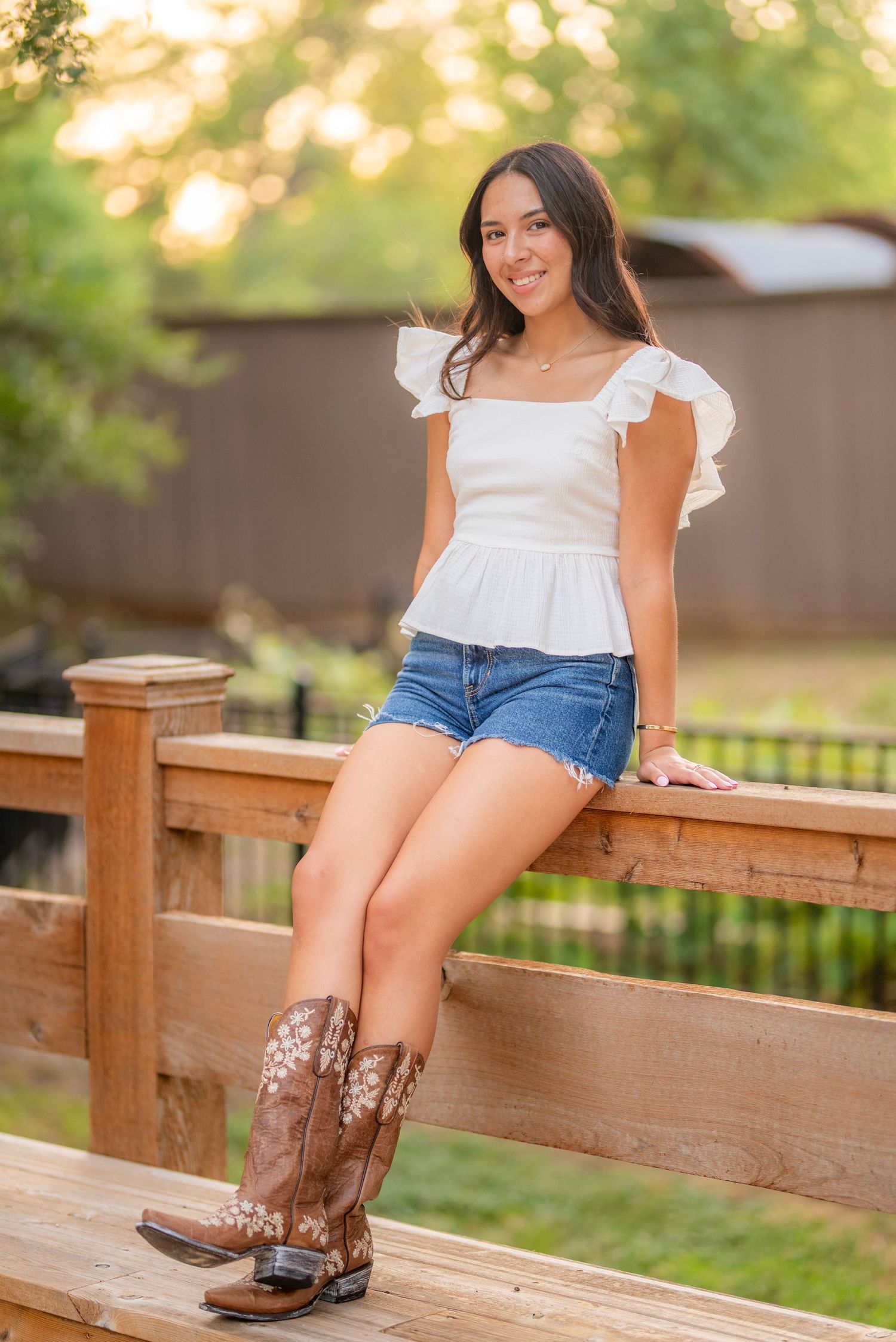 Young woman in white ruffled top, denim shorts, and brown cowboy boots sitting on a wooden fence outdoors.