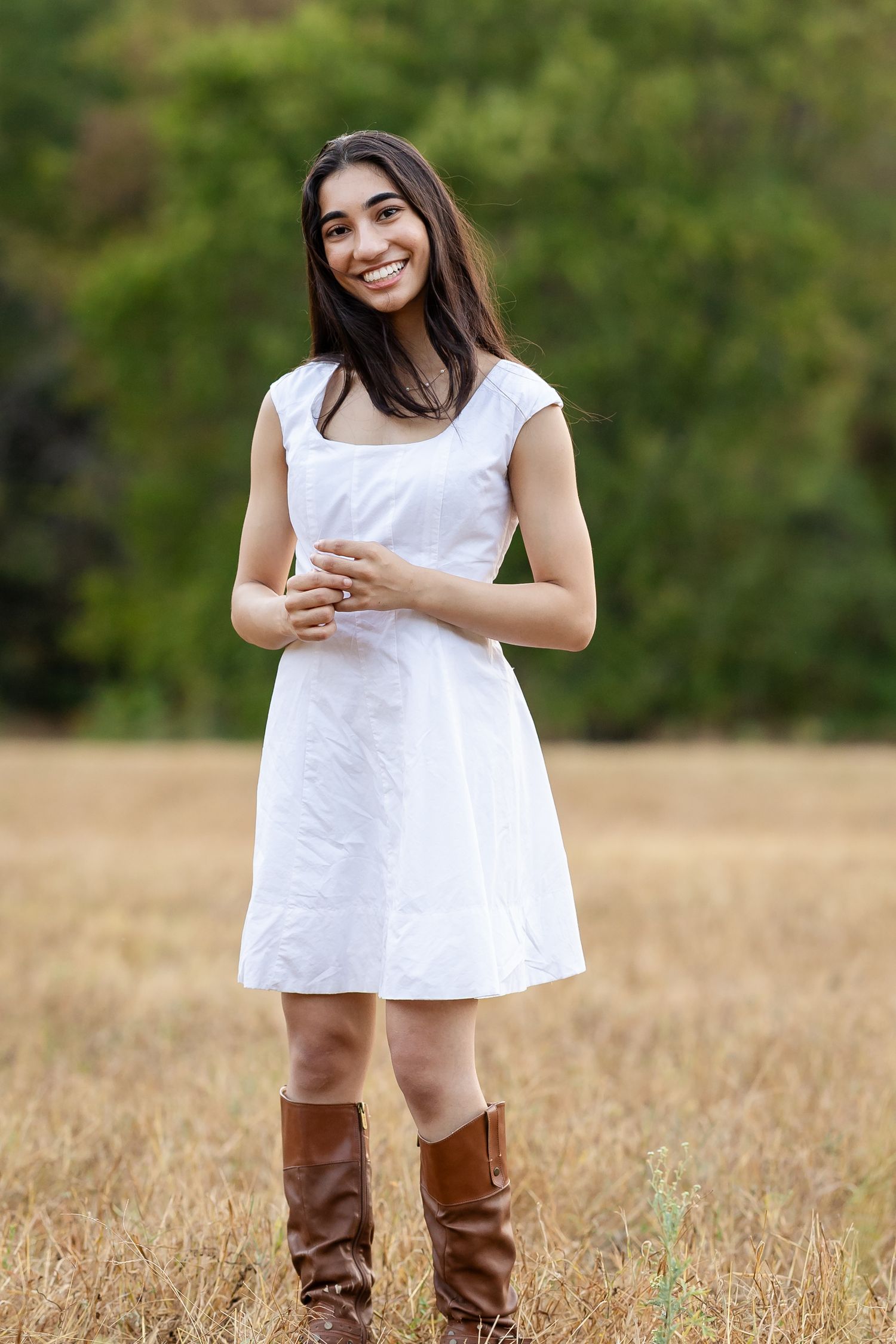 Young woman smiling in white dress and brown boots standing in golden field with green trees background.