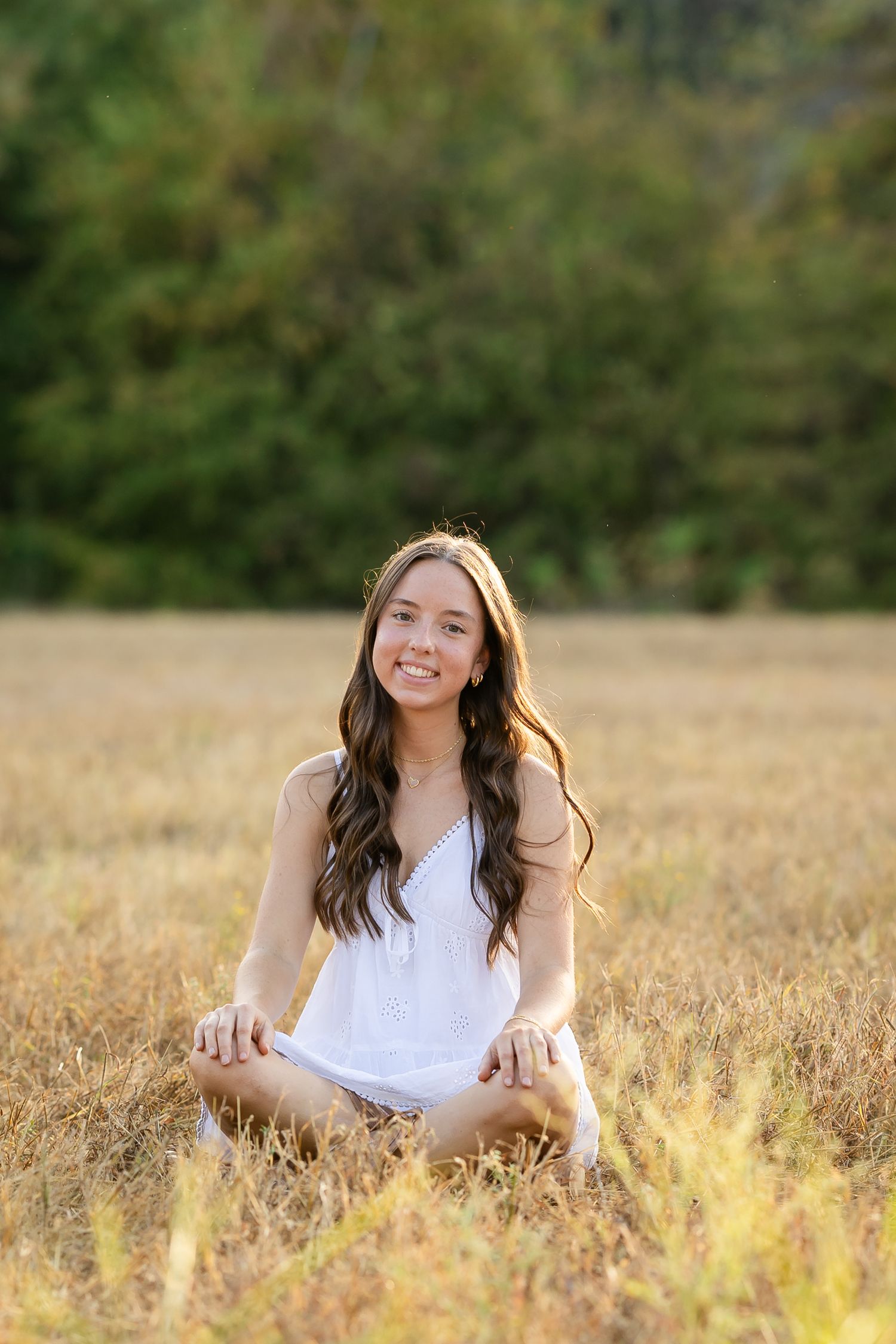 Young woman in white dress sitting cross-legged in golden field, smiling warmly with trees in background.