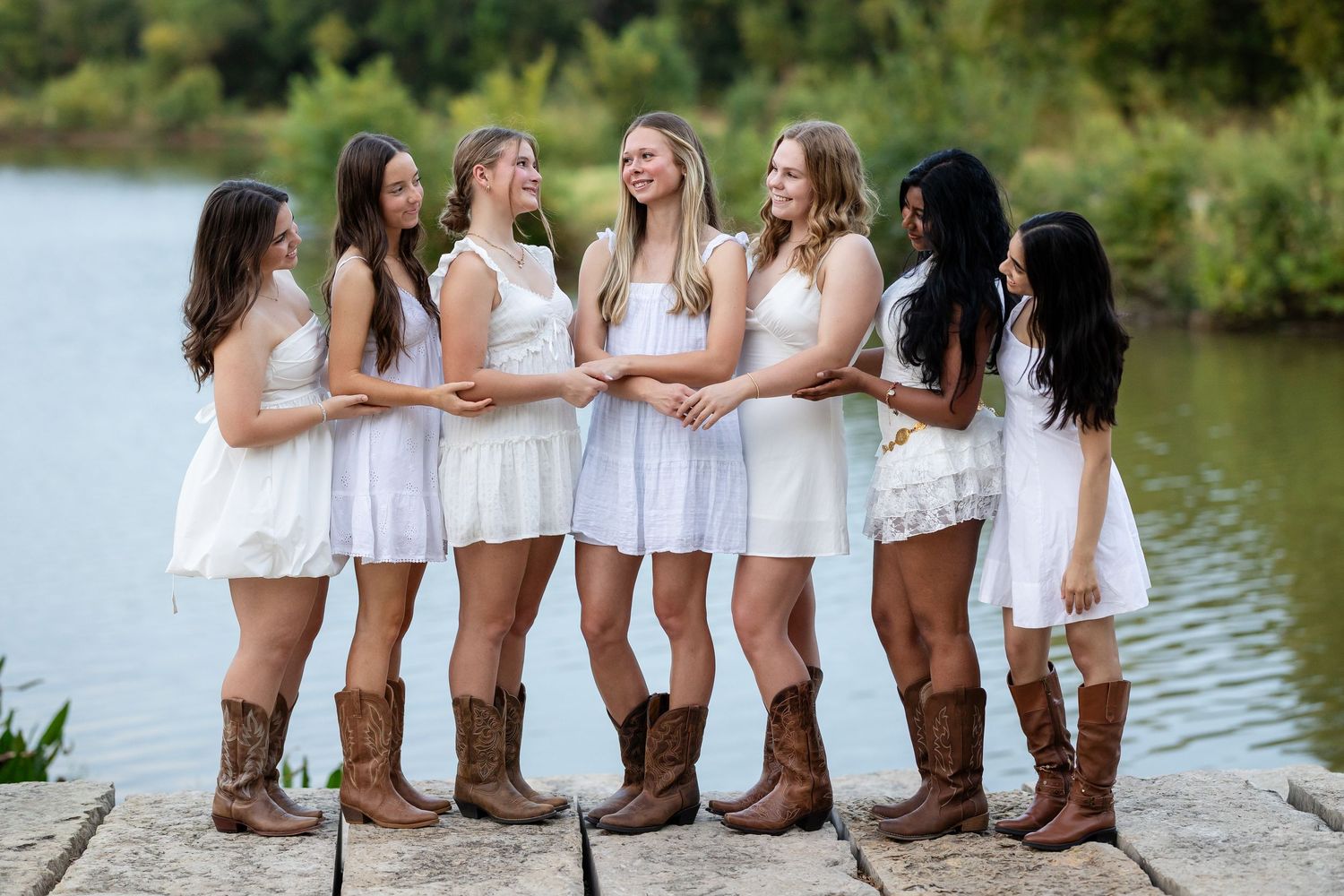 Seven young women in white dresses and brown cowboy boots pose together on a stone dock by a lake.