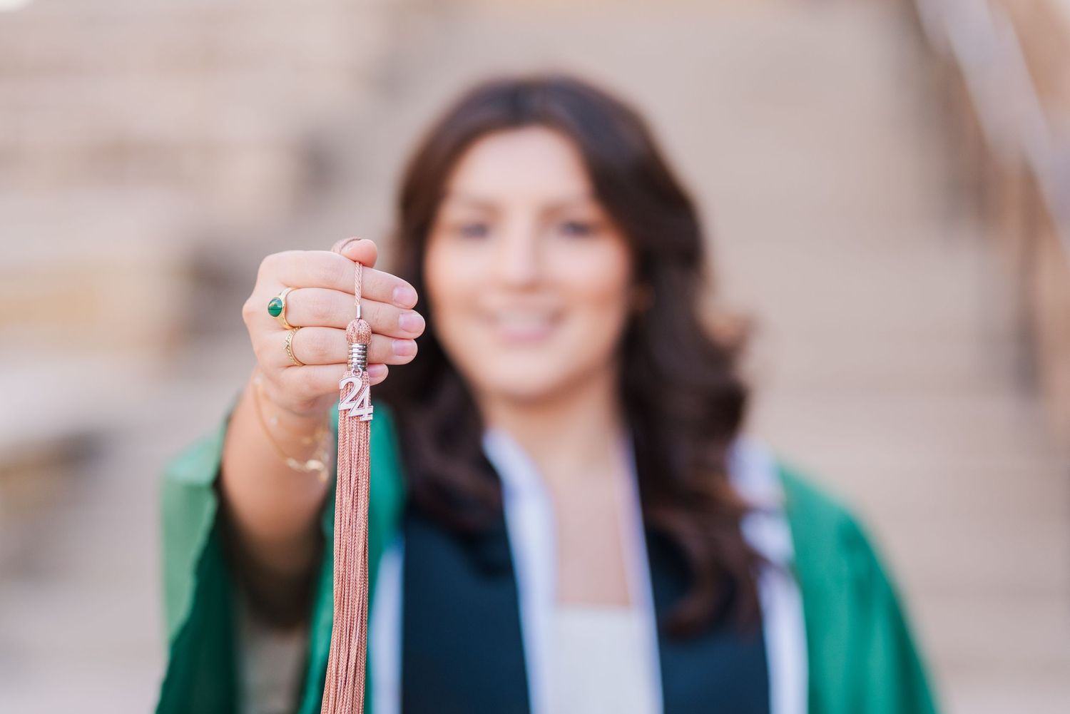 Smiling graduate in green gown holds colorful graduation cords toward camera on sunny steps.