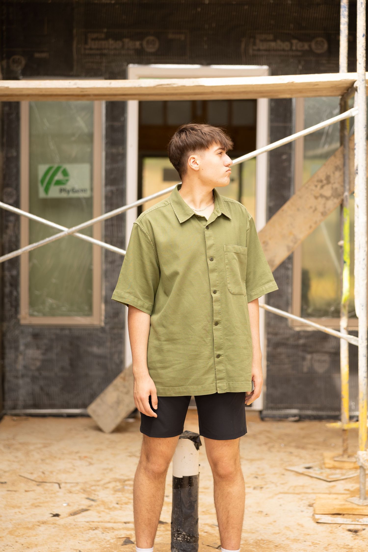 Young man in olive green short-sleeve shirt and black shorts standing at a construction site.