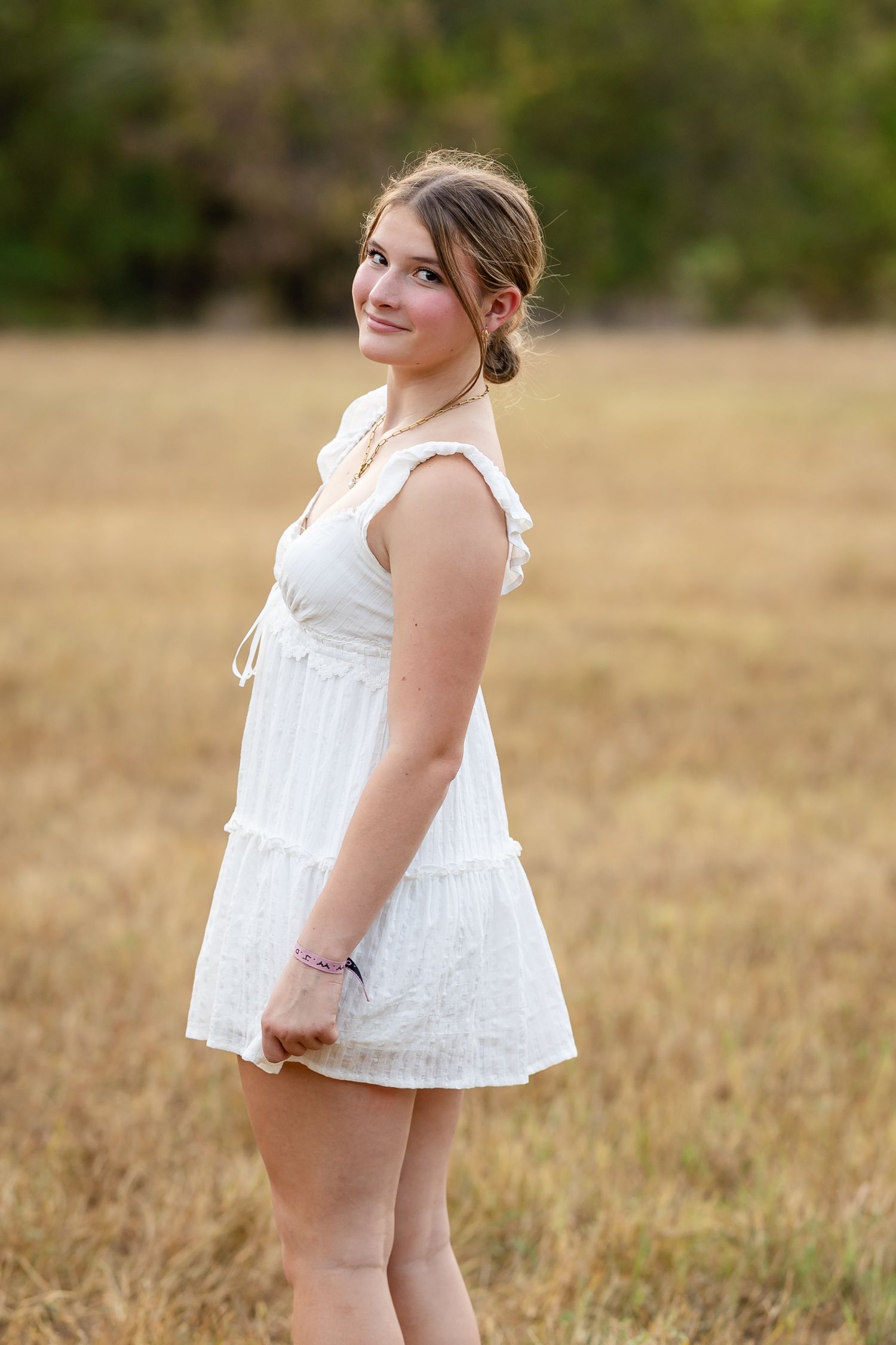 Young woman in white sundress smiling in golden field with trees in background.