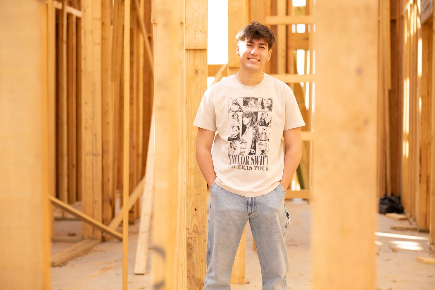 Young man in graphic tee and jeans smiling inside a wood-framed building under construction.