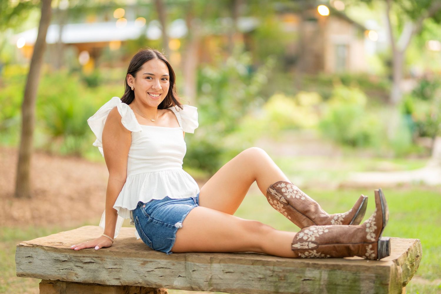 Young woman in white top, denim shorts, and cowboy boots sitting on a wooden bench outdoors.