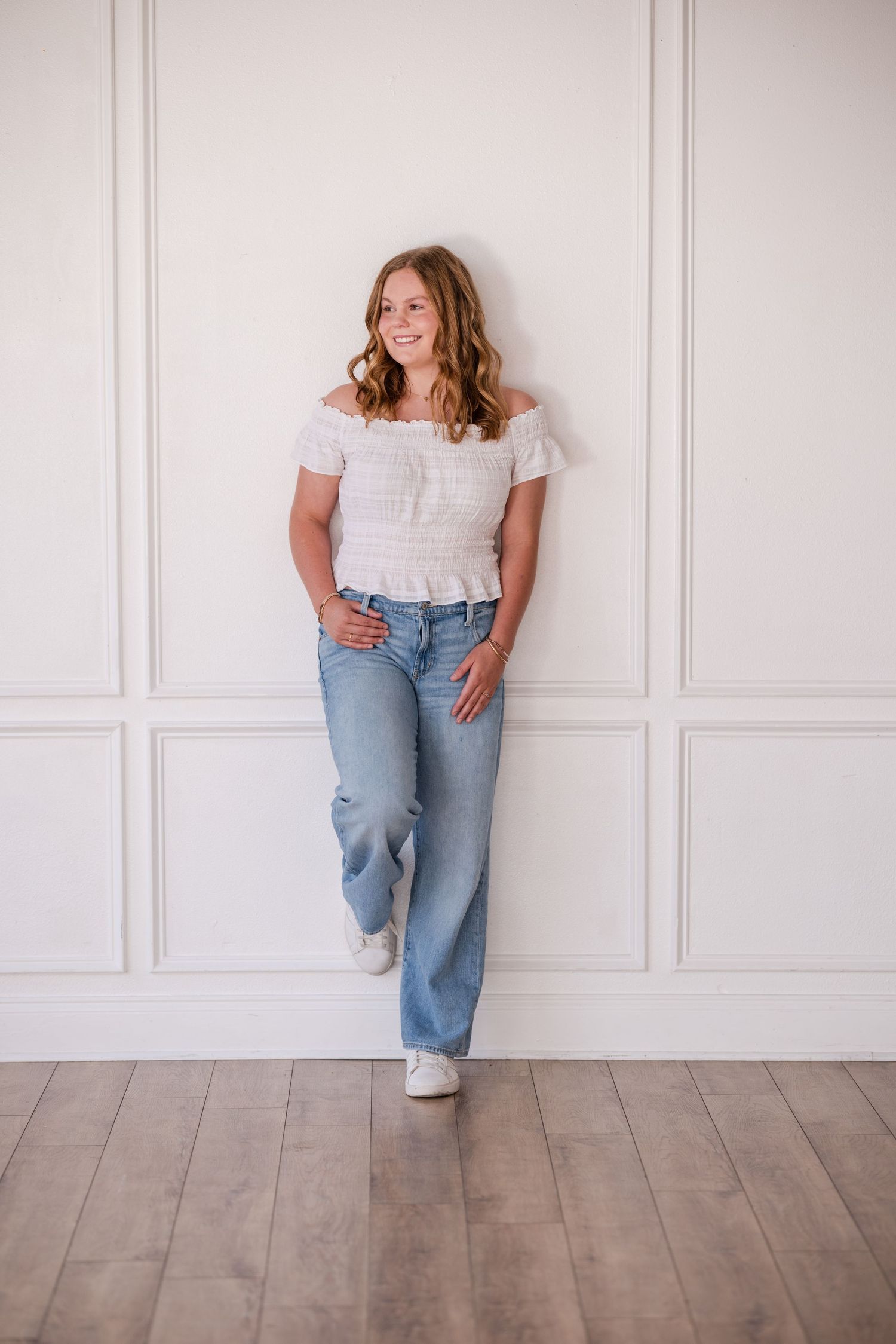 Smiling teenage girl in white off-shoulder top and blue jeans leaning against white paneled wall.