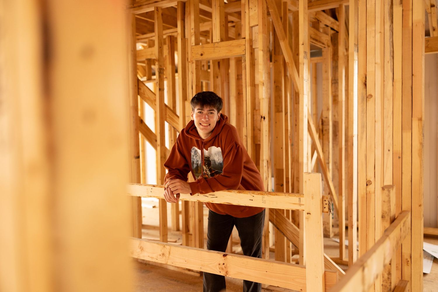 Young man smiling in an unfinished wood-framed house under construction, wearing an orange hoodie.