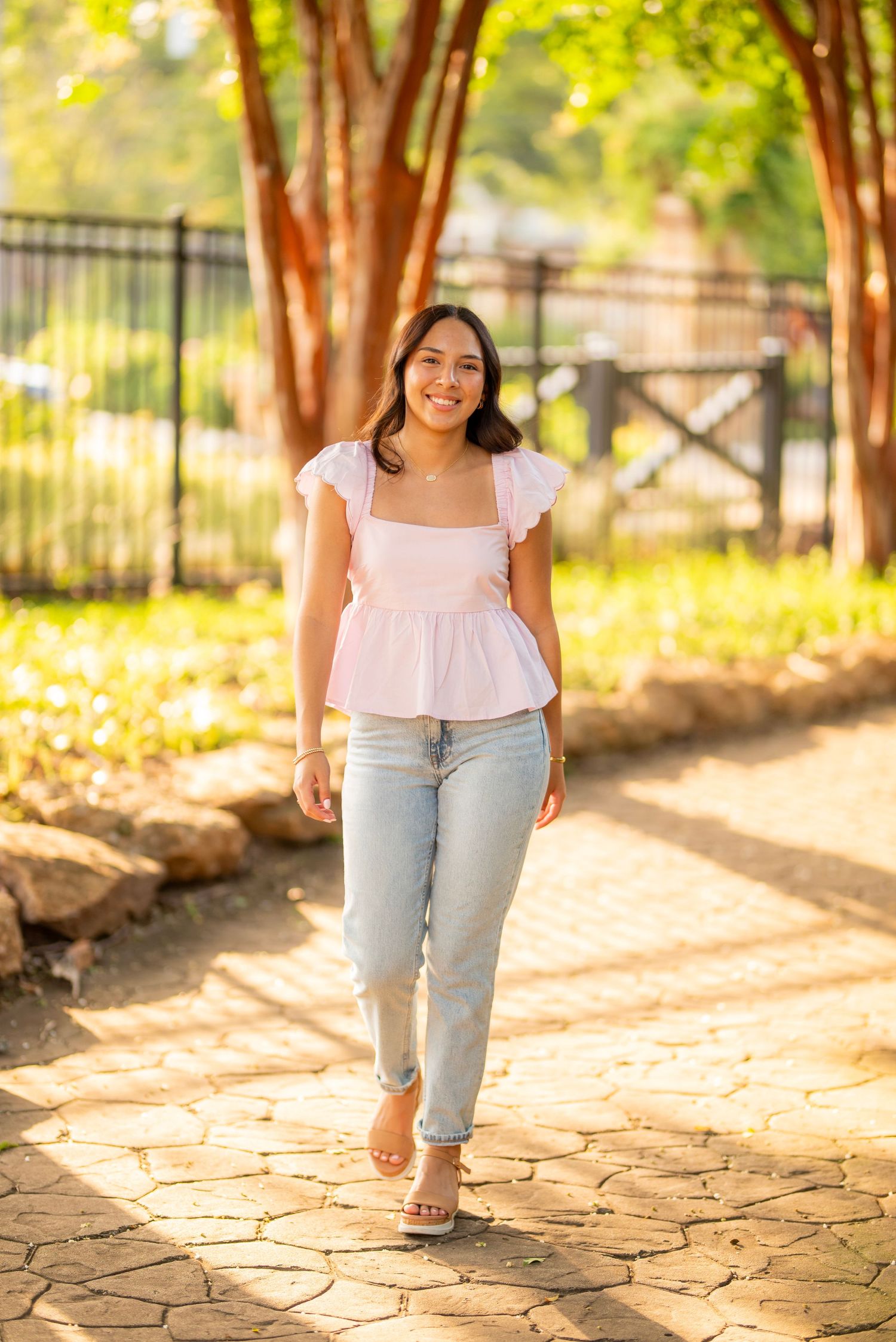 Young woman in pink peplum top and light jeans walks along a sunlit stone path surrounded by lush green trees.