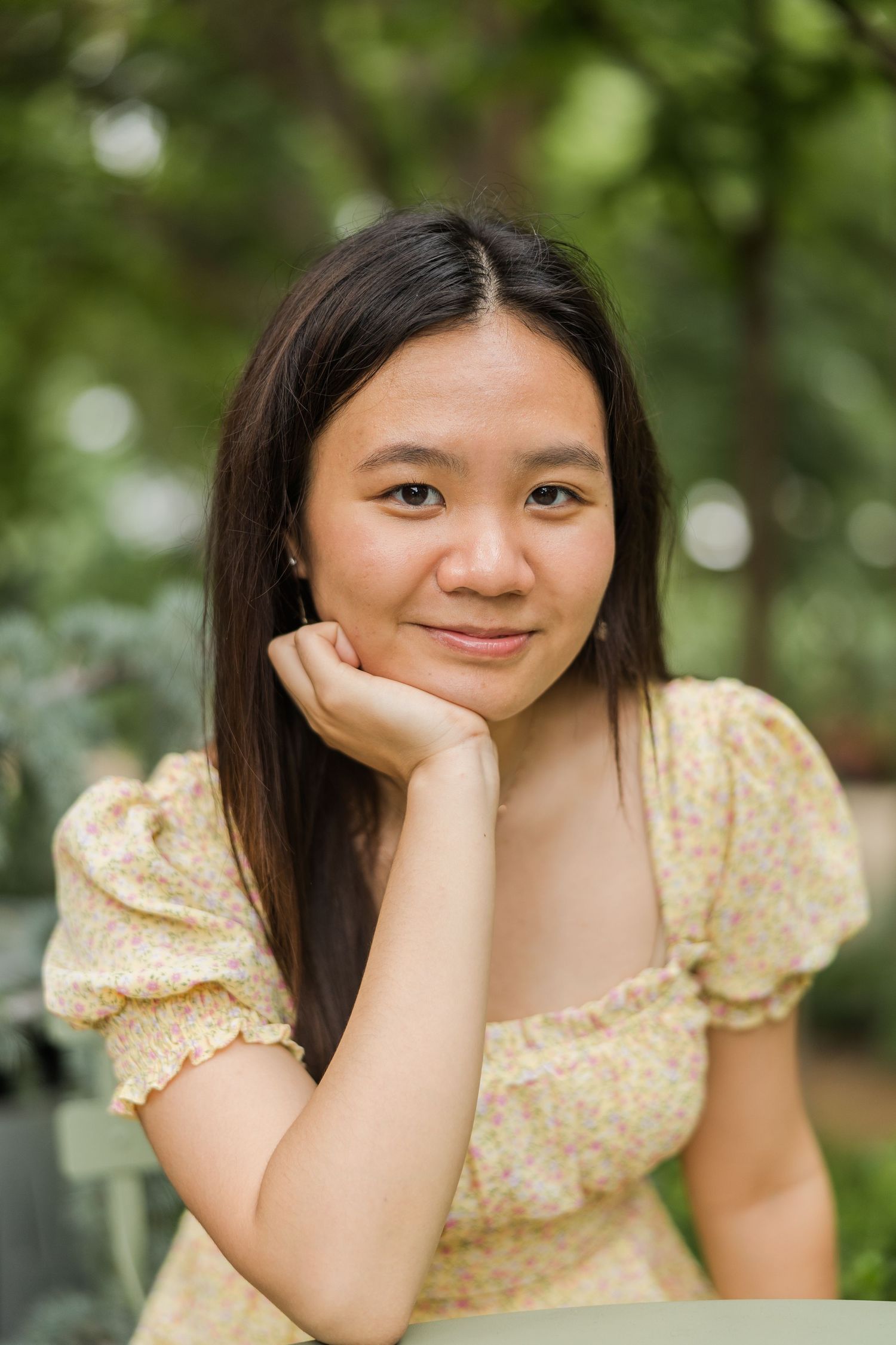 Young Asian woman in yellow floral dress resting chin on hand, smiling softly outdoors with green bokeh background.