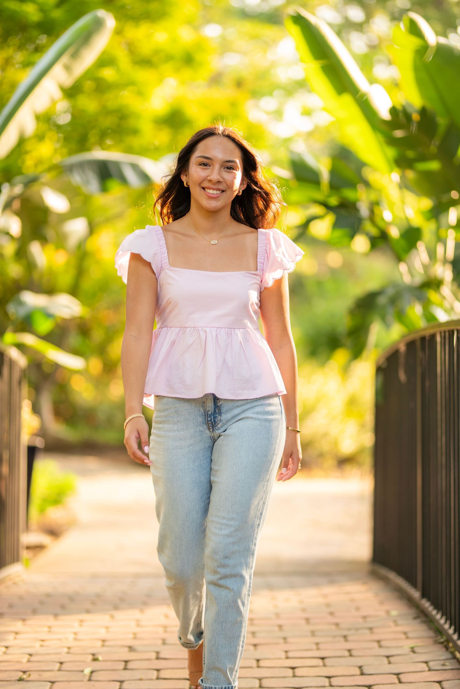 Young woman in pink peplum top and jeans walking on a brick bridge surrounded by lush green tropical foliage.