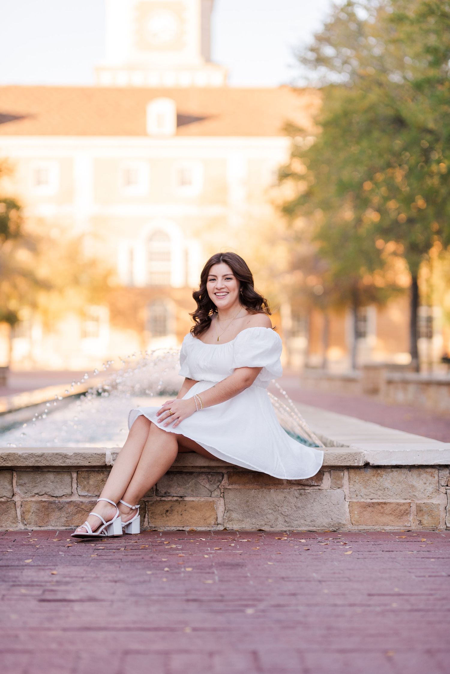 Young woman in white off-shoulder dress sits by fountain, smiling, wearing heels, with brick building backdrop.