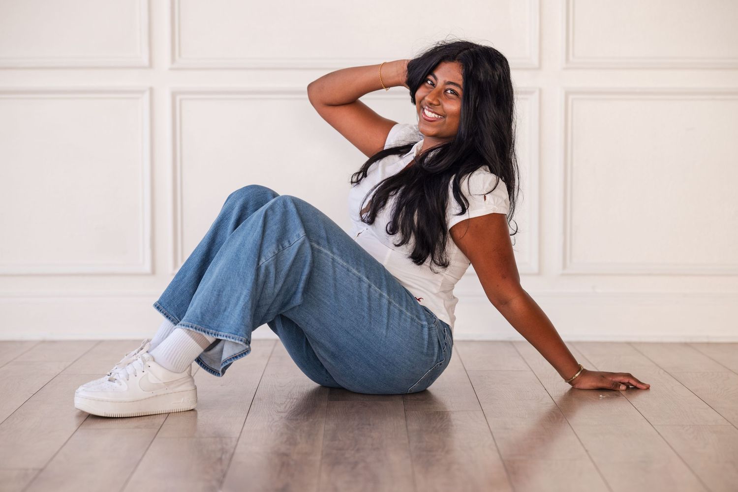 Young woman with long dark hair sits on hardwood floor smiling, wearing white tee, blue wide-leg jeans, and white sneakers.